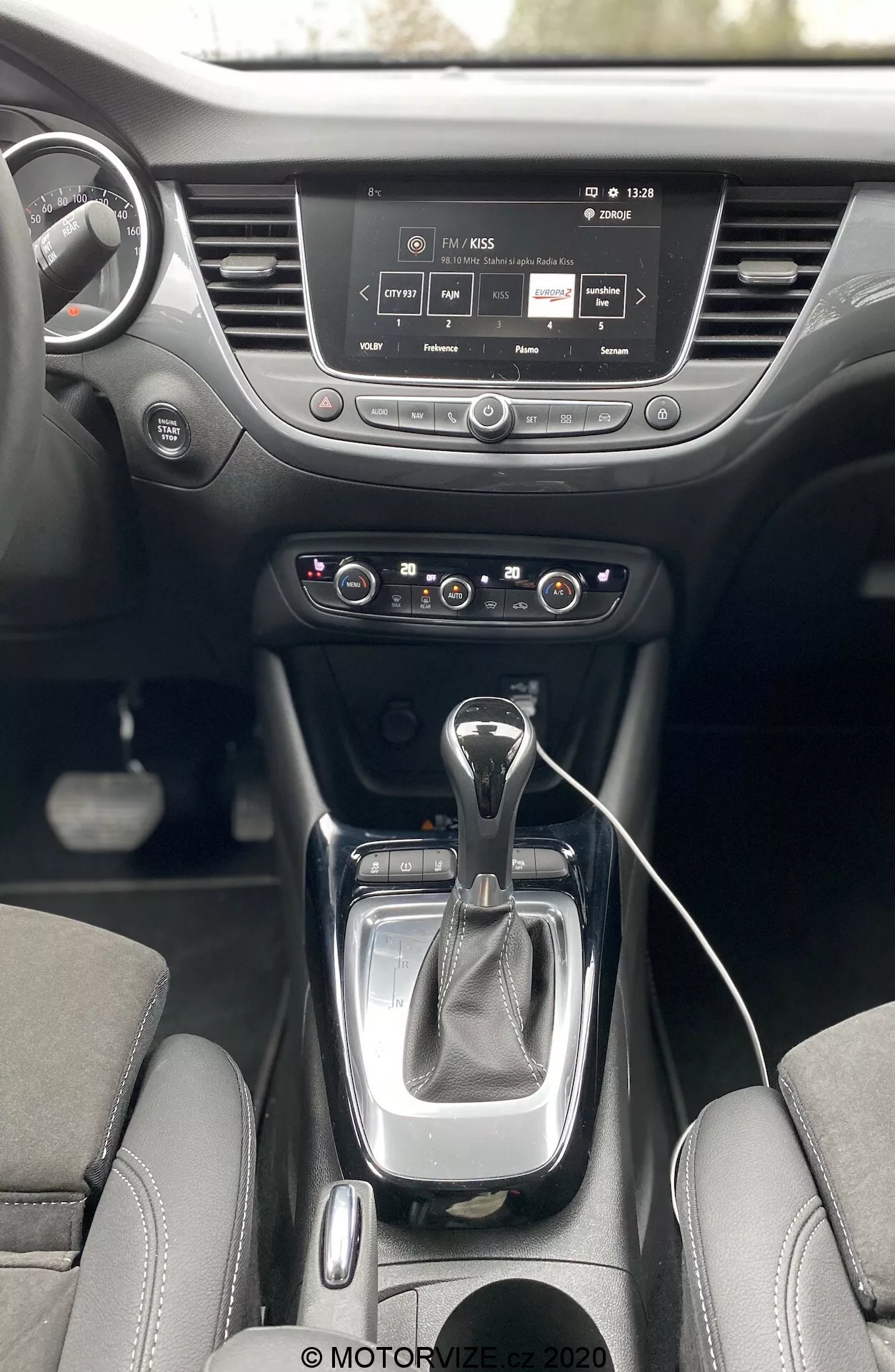 Interiér Opel Crossland 2021 z pohľadu prvej osoby, ukazujúci stredovú konzolu a prístrojovú dosku. Obsahuje modernú automatickú radiacu páku s koženým obalom, panel dvojzónovej klimatizácie, informačný a zábavný systém s dotykovým displejom, držiaky na nápoje a volant s ovládacími prvkami.