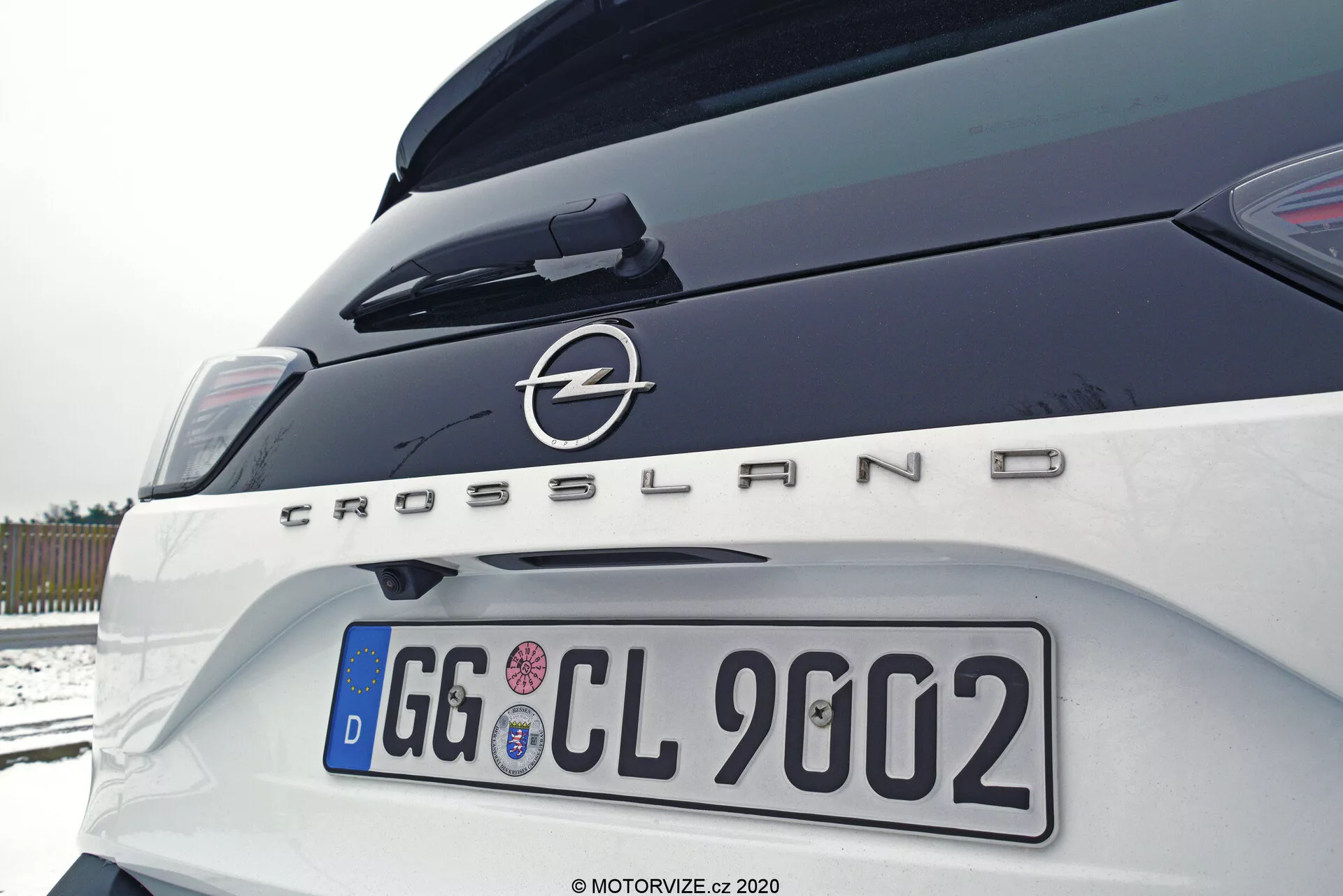 Detailný záber zadnej časti vozidla Opel Crossland 2021 z pravej strany v mierne nahor natočenom uhle, zobrazujúci kufor, pravé zadné svetlo, zadný nárazník a výrazné označenie značky a modelu s logom Opel a nápisom 'Crossland'. Európska ŠPZ a stopy snehu, naznačujúce vonkajšie, zamračené prostredie.