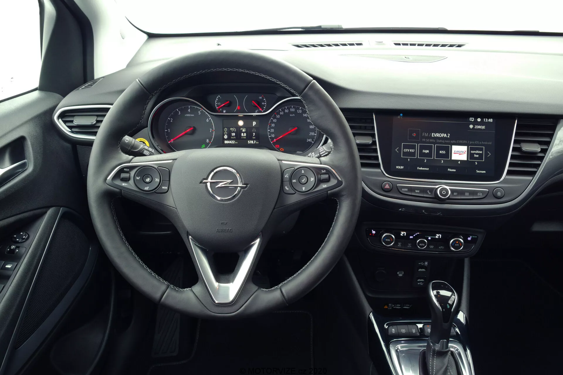 Interiér Opel Crossland 2021 z pohľadu sedadla spolujazdca, zobrazujúci kožený trojramenný volant s multifunkčnými tlačidlami, výrazné logo značky, analógový prístrojový panel s digitálnym displejom, stredovú konzolu s dotykovou obrazovkou infotainmentu, ovládače klímy, modernú automatickú páku a súčasný interiér s kombináciou materiálov, ako je koža, plast, chróm a kovové akcenty.