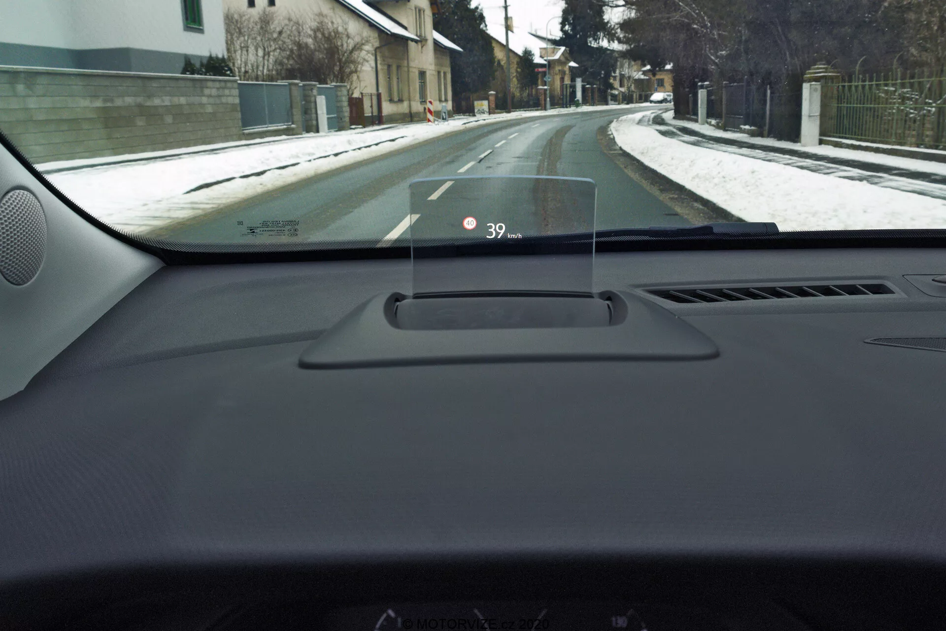 Pohľad z interiéru vozidla Opel Crossland 2021 z perspektívy vodiča, zvýrazňujúci Head-Up displej (HUD) na prístrojovej doske, zobrazujúci rýchlosť '39'. Pohľad zahŕňa prístrojovú dosku, okraj dverí na strane vodiča, A-stĺp a čiastočný pohľad na obytnú ulicu so snehom, čo naznačuje zimné podmienky.