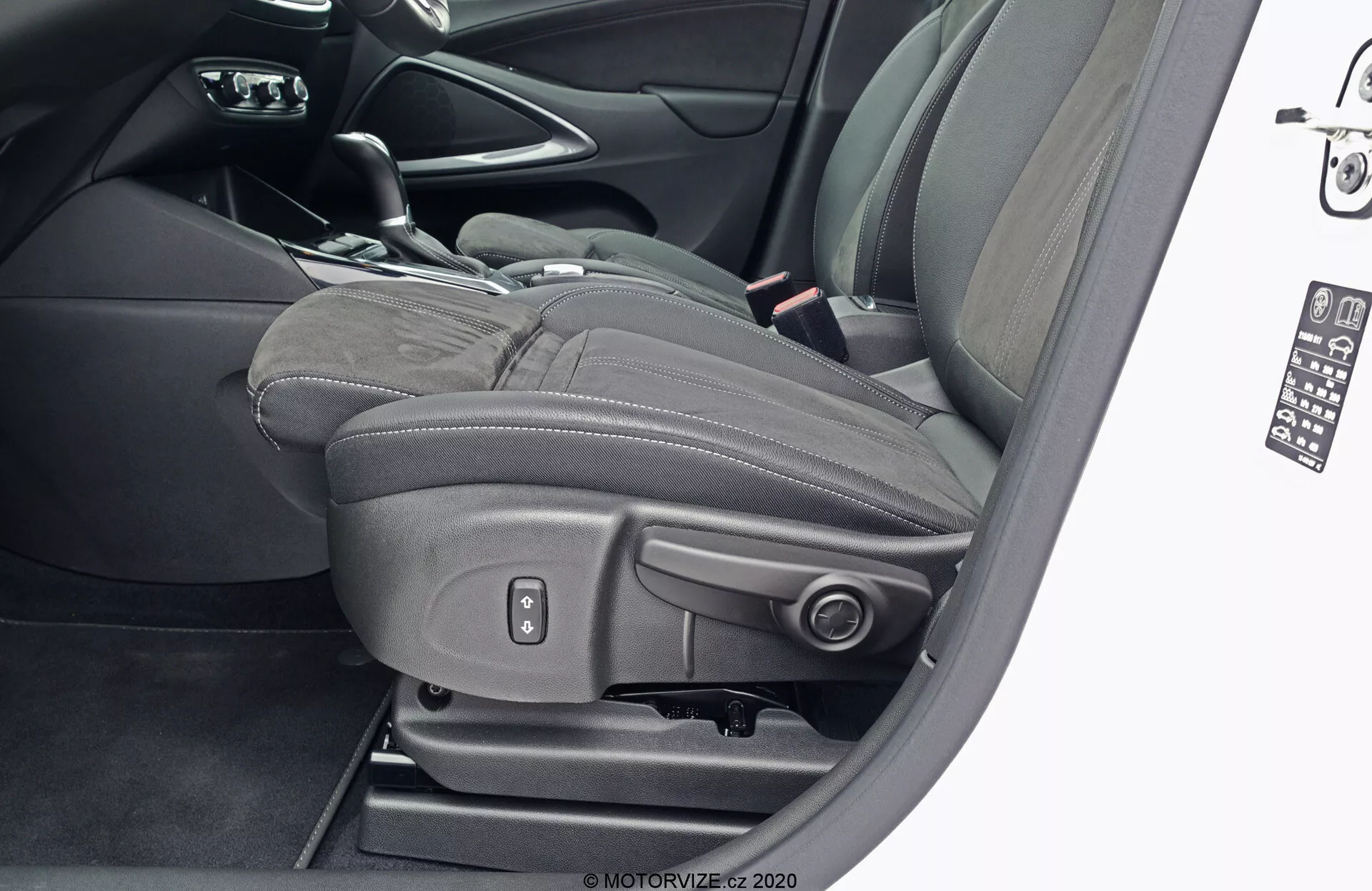 Detailný pohľad na sedadlo vodiča modelu Opel Crossland 2021 zvonku s otvorenými dverami, zobrazujúci čierne kožené čalúnenie s výrazným prešívaním a integrovanou nastaviteľnou opierkou hlavy, kovové ovládacie prvky pre elektrické nastavenie polohy sedadla, výšky, náklonu a bedrovej opierky, naznačujúce pokročilé komfortné a luxusné prvky.