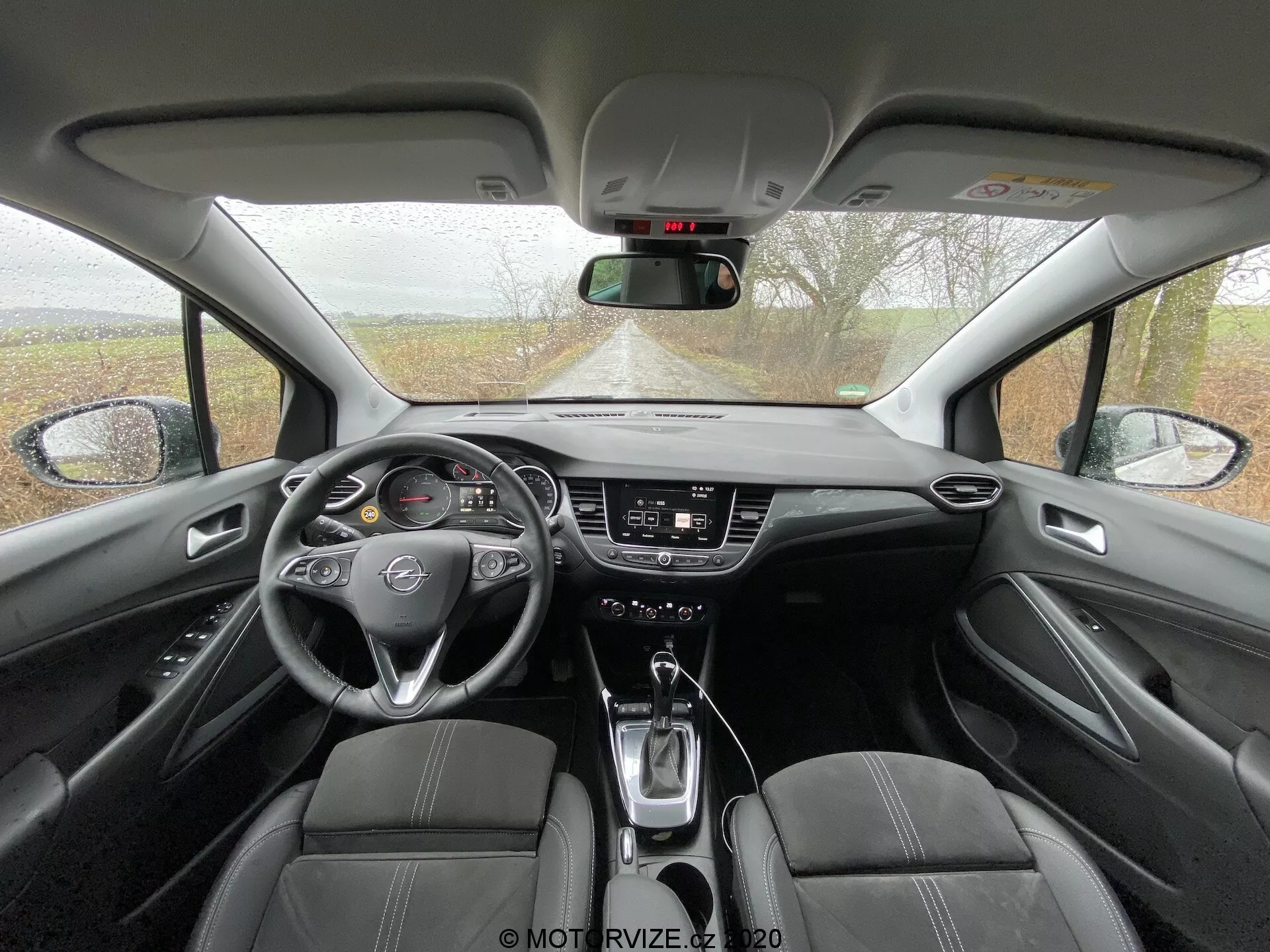 Výhľad do interiéru modelu Opel Crossland 2021 zo zadného sedadla, ktorý zobrazuje prednú palubnú dosku, trojramenný volant s emblémom, analógový a digitálny prístrojový panel, dotykový infotainment systém, ovládacie prvky klimatizácie, páku automatickej prevodovky, držiaky na nápoje, čalúnené predné sedadlá s perforovanými časťami, stredovú konzolu s opierkou, manuálne stmievateľné spätné zrkadlo, A-stĺpiky, dverové panely s opierkami a elektrickými ovládacími prvkami okien a mriežky reproduktorov zvukového systému.