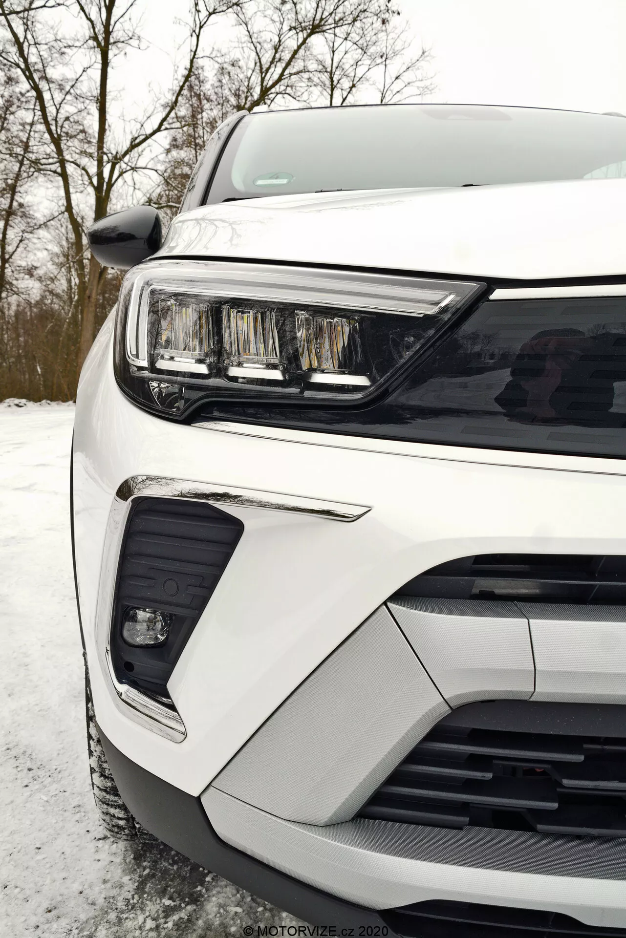 Detailný pohľad na prednú ľavú časť bieleho Opel Crossland 2021 fotografovaný z perspektívy úrovne zeme, zobrazený reflektor s elegantným vodorovným LED denným svetlom, časť mriežky s vodorovnými lamelami a predný nárazník s integrovaným hmlovým alebo prídavným jazdným svetlom v čiernom ráme a dolným výrezom, ktorý pravdepodobne slúži na prívod vzduchu alebo senzory pre asistenčné systémy vodiča.