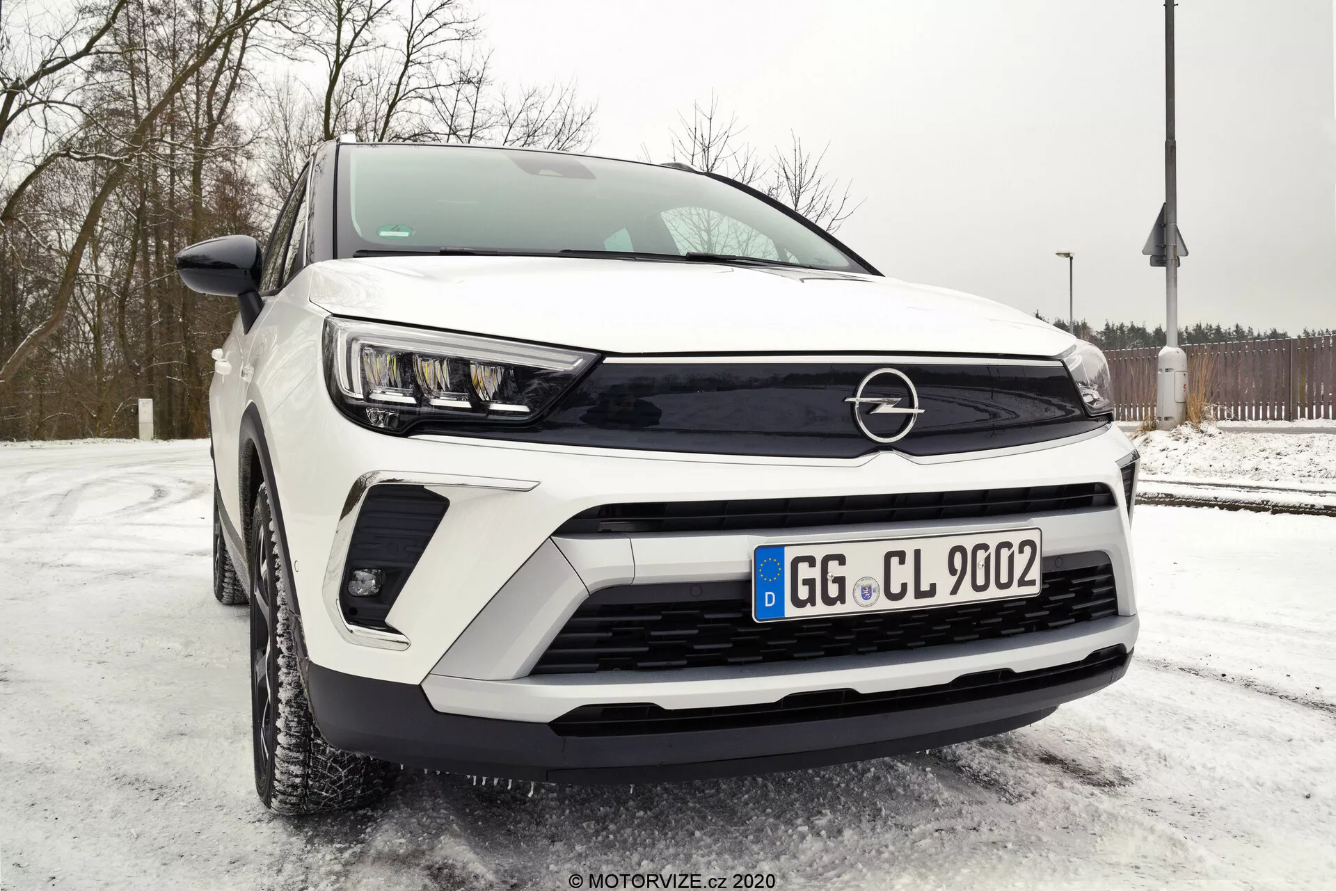 Nízky uhol pohľadu na SUV Opel Crossland 2021 so zameraním na pokročilé LED svetlomety, výraznú čiernu mriežku s emblémom, možné senzory asistenčných systémov vodiča a ľahkozliatinové disky kolies. Predná štvrtinová perspektíva zvýrazňuje ostré línie karosérie, chrániče nad kolesami a výrazný bočný profil z ľavej strany. Funkcie zahŕňajú vysoko technologické vonkajšie zrkadlá, čelné sklo so senzorom dažďa a zaparkovanie na zasneženom povrchu, čo naznačuje možný pohon všetkých kolies.