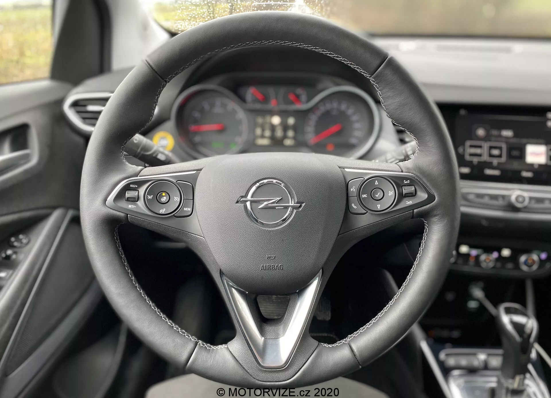 Interiér vozidla Opel Crossland 2021 z pohľadu vodiča, zvýrazňujúci trojramenný volant s multifunkčnými tlačidlami pre médiá, tempomat, hlasové príkazy a telefón. Logo Opel a označenie airbagu sú jasne viditeľné. Za volantom je čiastočne viditeľný analógový prístrojový panel so stredovým rýchlomerom, otáčkomerom a ďalšími meračmi. Palubná doska ukazuje rozmazané tlačidlá a možnú centrálnu dotykovú obrazovku pre infotainment systém.