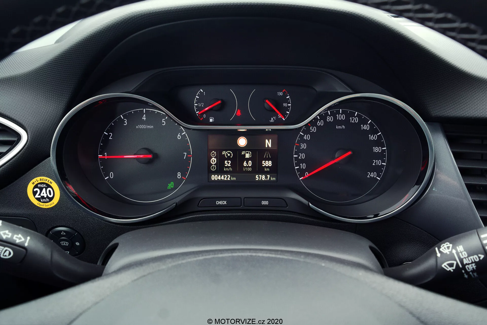 Detailný pohľad na prístrojovú dosku a časti volantu modelu Opel Crossland 2021. Panel obsahuje dva hlavné analógové ciferníky pre otáčkomer a rýchlomer, s červenými ukazovateľmi, a centrálny digitálny displej zobrazujúci aktuálnu rýchlosť, počet najazdených kilometrov, počet najazdených kilometrov na aktuálnej ceste a výstražné ikony. Snímka tiež ukazuje ovládacie páčky po stranách volantu, čo naznačuje technologickú integráciu v palubnej doske vozidla.