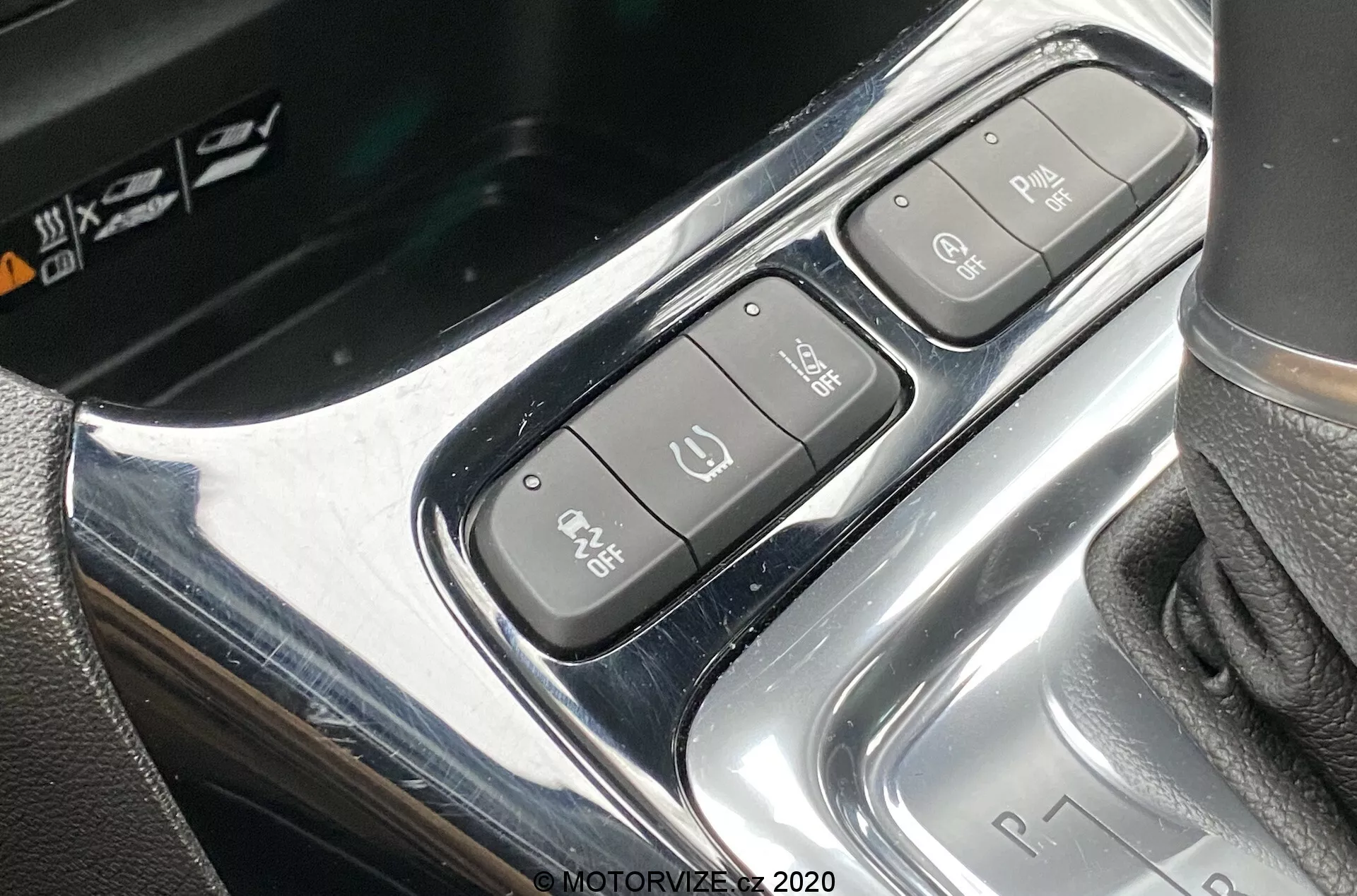 Detailný záber na stredovú konzolu Opel Crossland 2021, zameraný na ovládací panel v blízkosti radiacej páky. Záber zhora a mierne pod uhlom ukazuje chrómovaný dizajn s tromi rôznymi tlačidlami: elektronická parkovacia brzda, Auto Hold a parkovací asistent. Radiaca páka napravo zobrazuje sekvenciu 'P R N D', čo značí automatickú prevodovku.
