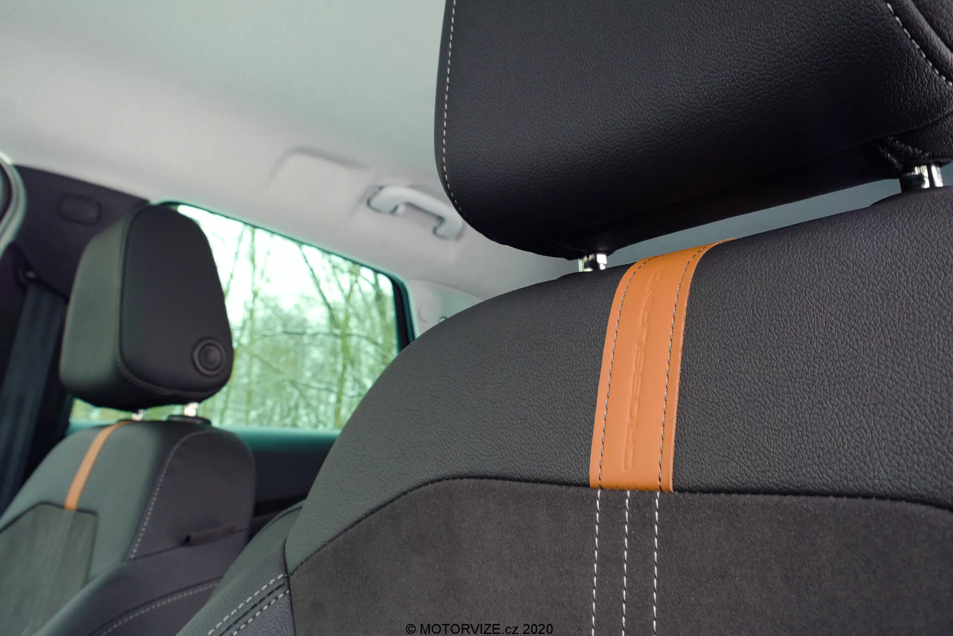 Detailný pohľad na interiér Opel Crossland 2021, zameriavajúci sa na predné a zadné sedadlá. Obrázok je zachytený z perspektívy zadného sedadla, mierne dopredu a doľava, ukazujúc opierku chrbta predného sedadla spolujazdca a časť sedadla vodiča. Sedadlá majú tmavé čalúnenie s výrazným oranžovým prešívaním, ktoré naznačuje športovú tému, a integrované opierky hlavy naznačujúce moderný dizajn. Bočný výstuž predného sedadla spolujazdca zvýrazňuje športovosť a dodatočnú bočnú podporu. Prirodzené svetlo prechádzajúce cez bočné okná a viditeľný B-stĺpik zdôrazňujú estetiku interiéru, zatiaľ čo vodoznak '© MOTORVIZE.cz 2020' v pravom hornom rohu naznačuje pôvod obrázku.