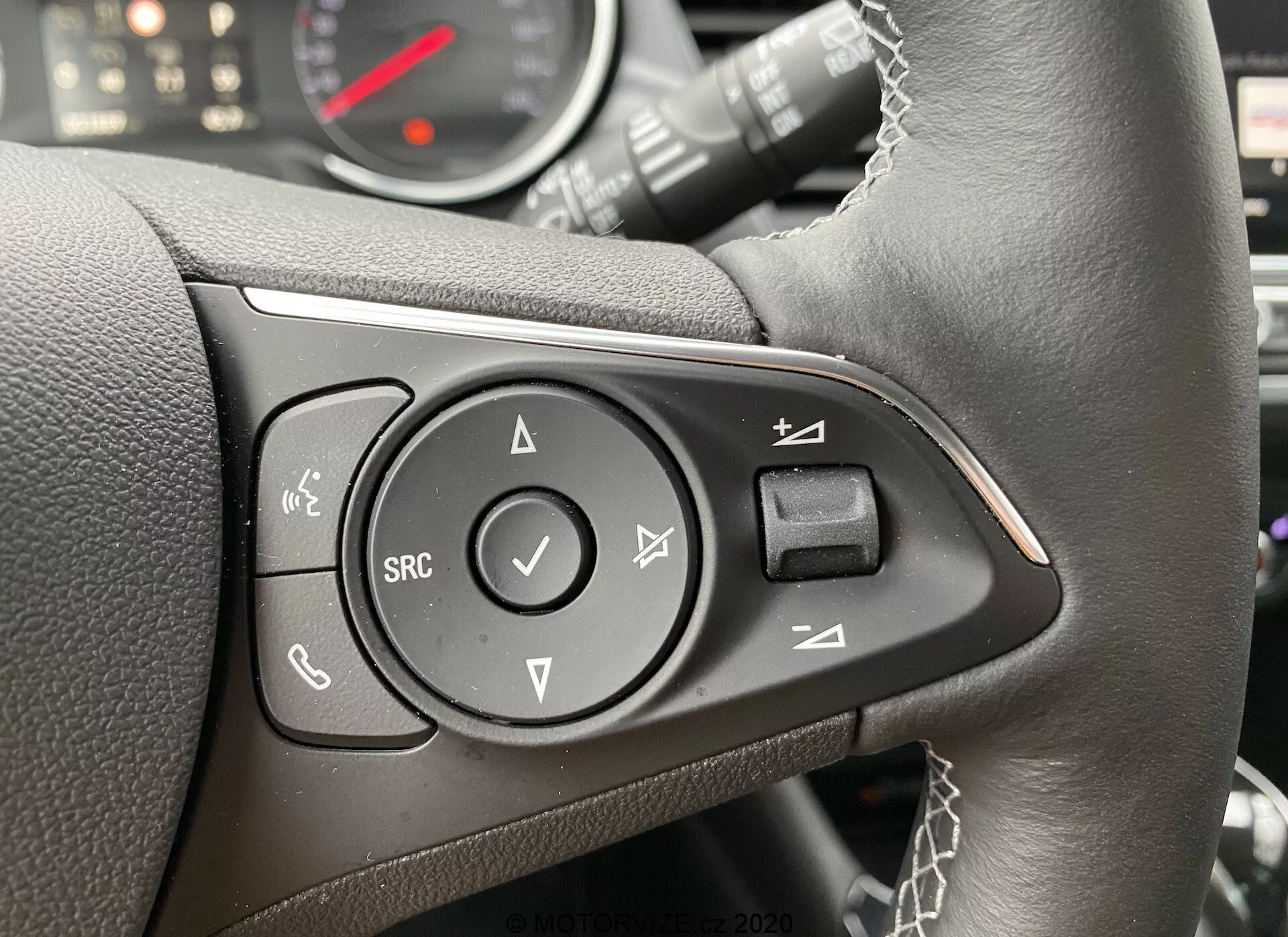 Detailný záber na multifunkčné ovládacie prvky volantu Opel Crossland 2021 z pohľadu vodiča, mierne posunutý doprava. Čierny kožený volant s viditeľným šitím a ovládacími prvkami umiestnenými v lesklom čiernom paneli. Ľavé tlačidlo je určené na hands-free volania, pravdepodobne s podporou Bluetooth. Nad ním sa nachádza tlačidlo na ovládanie hlasom. Stredné smerové pole so šípkami hore, dole, vľavo a vpravo a tlačidlom 'OK'. Pod poľom je tlačidlo 'SRC' na prepínanie zvukových zdrojov. Prístrojový panel, vrátane tachometra a pravdepodobne aj otáčkomera, je čiastočne viditeľný a rozmazaný.