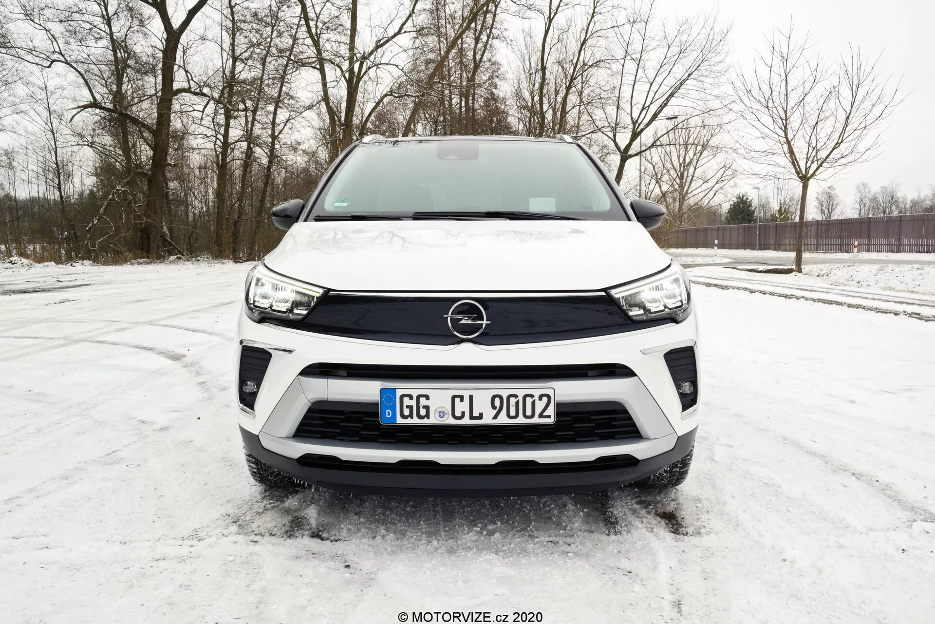 Čelný pohľad na moderné biele SUV Opel Crossland 2021 zaparkované na zasneženej ploche, s nízkoúhlovým záberom zdôrazňujúcim prednú mriežku, dizajn svetlometov a čierne detaily nárazníka. Kľúčové prvky zahŕňajú výrazné logo Opel, elegantné LED svetlomety, štandardné umiestnenie evidenčnej tabuľky, potenciálnu integráciu parkovacích senzorov a robustný dizajn nárazníka naznačujúci schopnosť jazdy v teréne.
