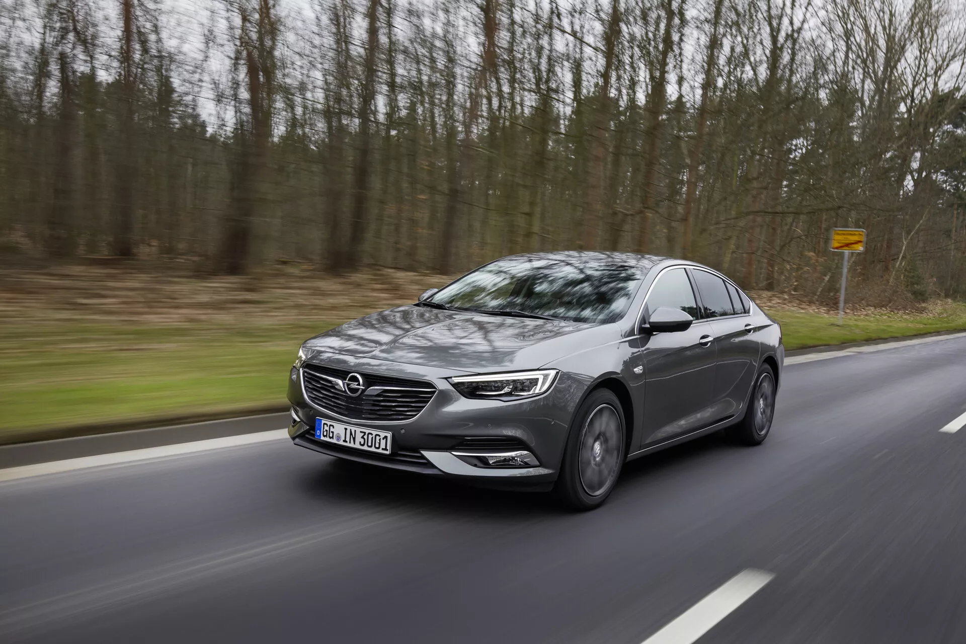 Moderný sedan Opel Insignia 2013 v pohybe na kľukatej ceste cez les, zobrazujúci bočný profil s LED svetlometmi, veľkou mriežkou, zliatinovými kolesami, bočnými zrkadlami s ukazovateľmi a tónovanými oknami