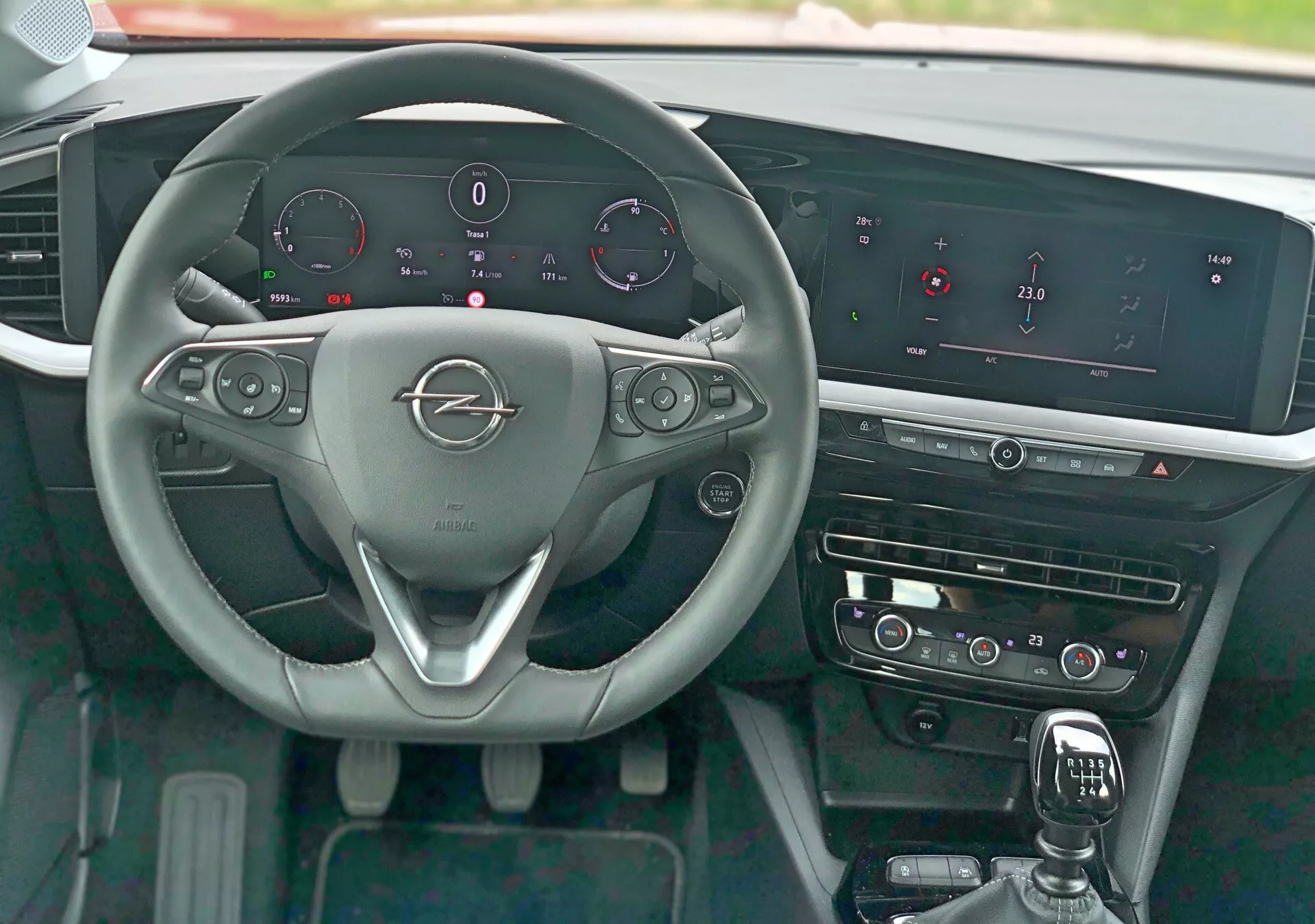 Pohľad z interiéru Opel Mokka 2021 z pohľadu vodiča ukazuje trojramenný volant s kovovým logom značky, ovládacie prvky volantu pre audio, telefón a tempomat, pádla pre radenie, digitálny prístrojový panel zobrazujúci rýchlosť, otáčky, hladinu paliva a teplotu motora, výrazný infotainment systém so širokou horizontálnou obrazovkou, dotykové ovládanie HVAC, radiacu páku s koženým poťahom, leštené ozdobné prvky a tlačidlo bezkľúčového štartu.
