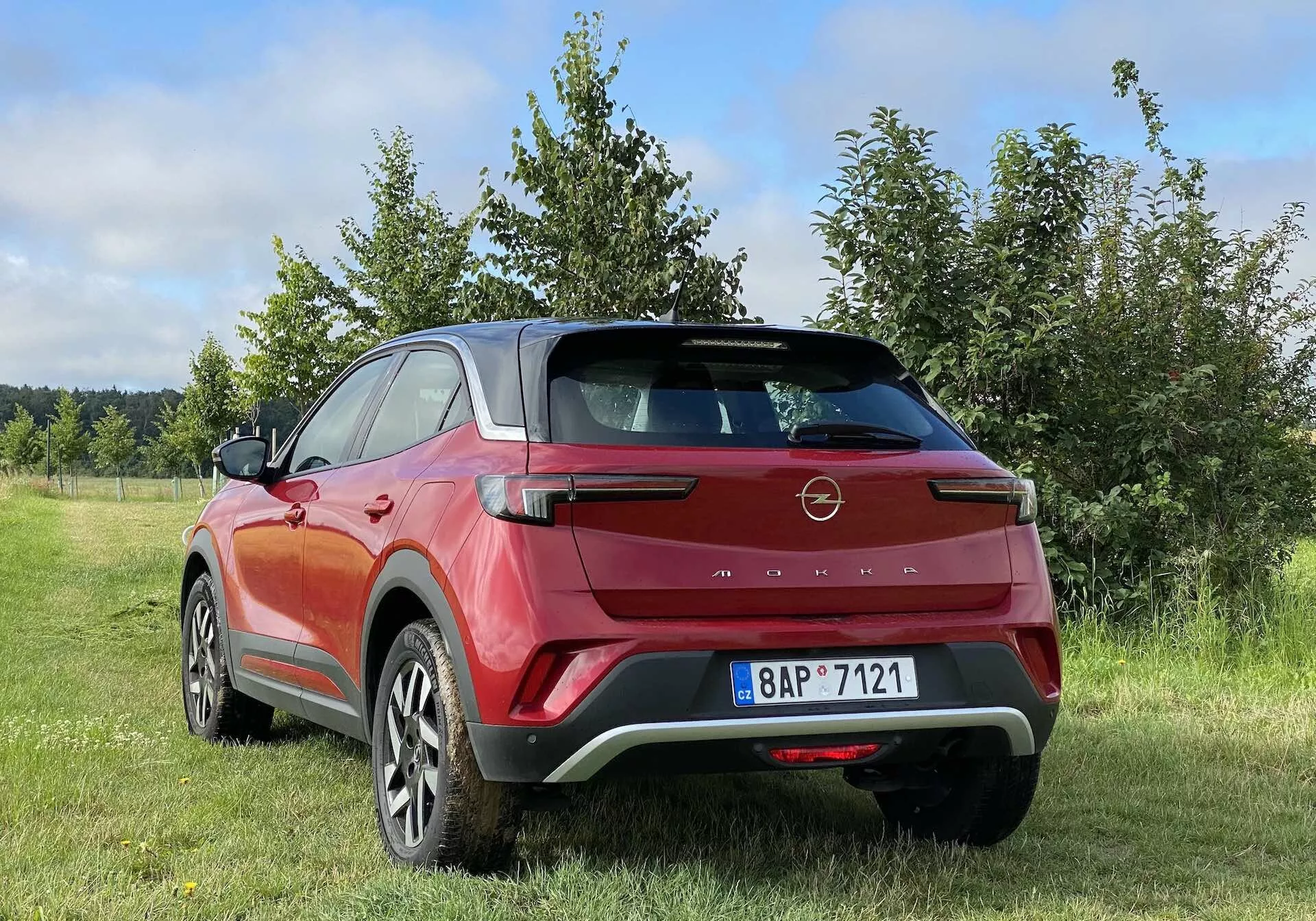 Farebná fotografia zobrazujúca zadný trojštvrťový pohľad na moderný crossover SUV Opel Mokka 2021 v červeno-čiernej farbe, zaparkovaný vonku na trávnatom pozemku so stromami alebo kríkmi v pozadí. Dizajnové prvky vozidla zahŕňajú aerodynamický zadný spoiler, hranaté LED zadné svetlá, vysoko posadenú brzdovú lampu, dvojfarebnú karosériu, veľké zliatinové kolesá, zapustené kľučky dverí a čierne plastové opláštenie. Fotografia je zhotovená z nízkeho uhla, zdôrazňujúc výšku a svetlosť SUV, s čiastočne zamračenou oblohou, naznačujúcou premenlivé poveternostné podmienky.