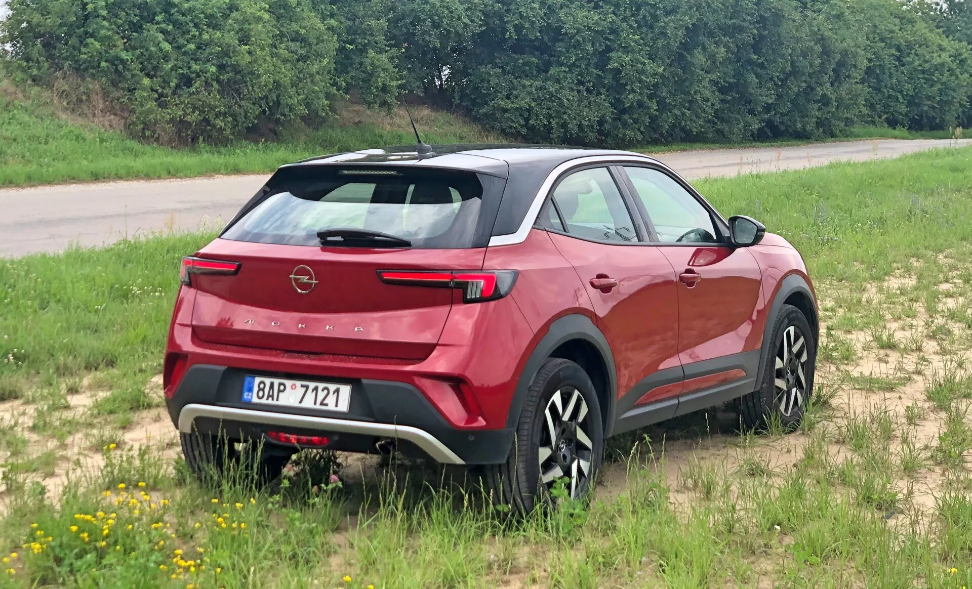 Moderné crossover SUV Opel Mokka 2021 zachytené z trojštvrťovej zadnej perspektívy, zvýrazňujúce zadný spojler, ostrý zadný sklo, viditeľný znak značky a elegantné zadné svetlá. Vozidlo je umiestnené na trávnatom povrchu, čo poukazuje na jeho ľahkú terénnu schopnosť. Bočný profil ukazuje tvarované línie karosérie, čierne obklady a štýlové viacramenné disky. Integrovaný zadný stierač, parkovacie senzory a kontrastná ochranná doska naznačujú aerodynamiku, praktickosť a robustnosť.