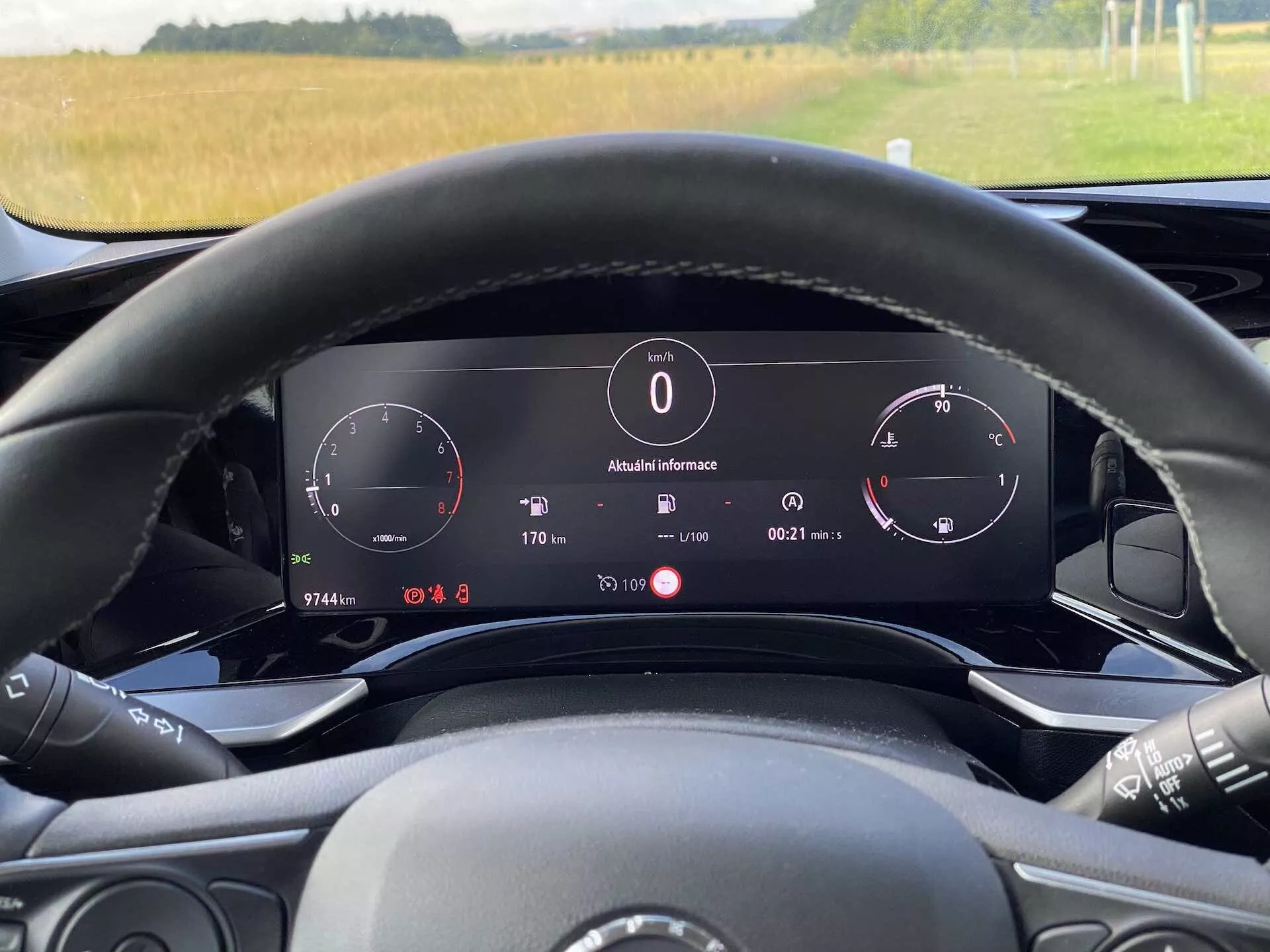 Interiérový pohľad na Opel Mokka 1.2 DI Turbo 2021 z pohľadu vodiča za volantom. Obrázok zdôrazňuje digitálnu prístrojovú dosku za volantom, ktorá zahŕňa rýchlomer, tachometer a informačný panel zobrazujúci vonkajšiu teplotu (8°C), hodiny, polohu prevodovky ('P' pre parkovanie) a podrobnosti palubného počítača. Volant obsahuje viditeľné ovládacie prvky pre médiá a je čiastočne viditeľný. Prirodzené osvetlenie naznačuje dennú dobu a cez čelné sklo je vidieť vonkajší výhľad na pole. Vozidlo ukazuje elektronickú zobrazovaciu palubnú dosku a pokročilé technologické funkcie.