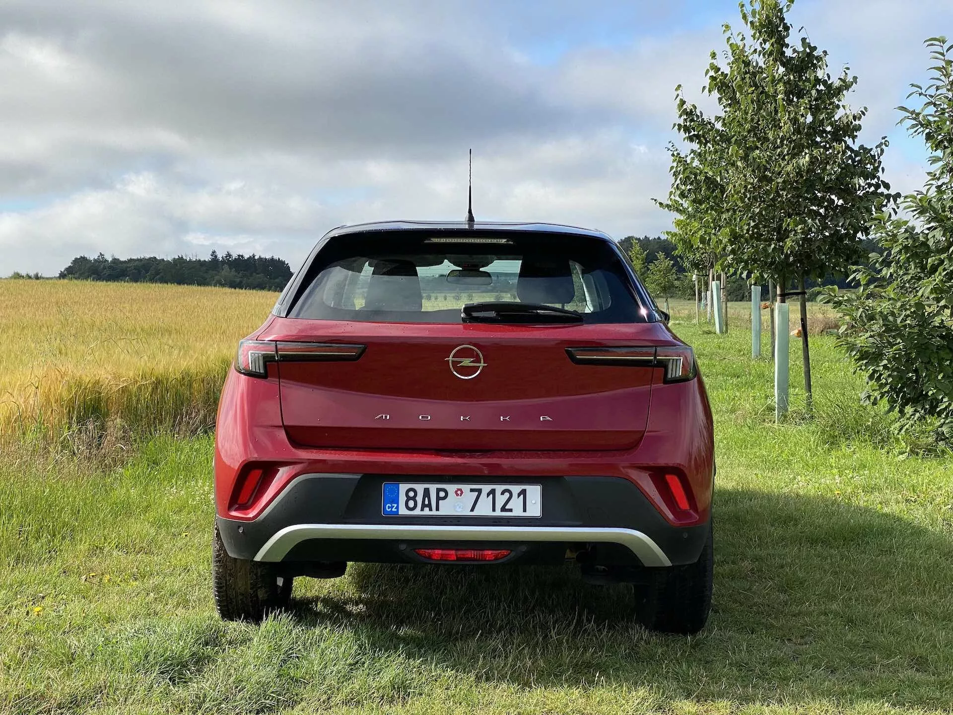 Zadný pohľad na červený Opel Mokka 1.2 DI Turbo 2021, vyznačujúci sa moderným dizajnom s ostrými líniami a emblémom výrobcu na zadnom veku, charakteristickými zadnými LED svetlami, zadným nárazníkom s kontrastnými vložkami, parkovacími senzormi, integrovanými reflektormi, stieračom zadného okna, anténou v tvare žraločej plutvy, zadným spojlerom a registračnou značkou v rurálnom prostredí s trávou, poľom a stromami.