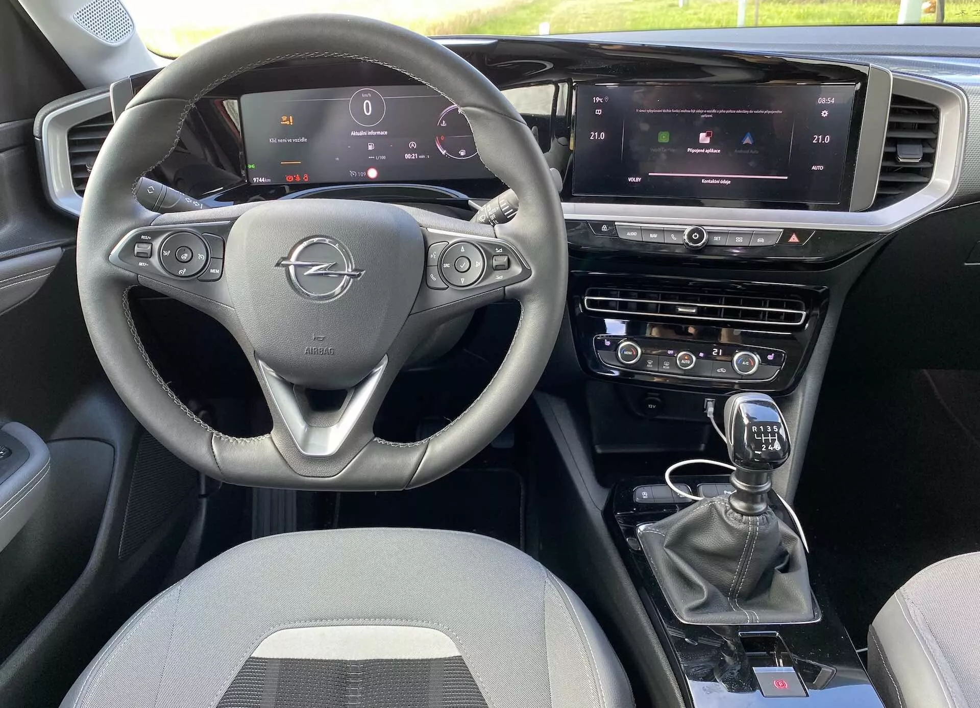 Interiér Opel Mokka 2021 z pohľadu predného spolujazdca, trojramenný volant s ovládacími prvkami, plne digitálny prístrojový panel, centrálny dotykový displej infotainmentu s ovládaním klímy a navigáciou a manuálna radiaca páka. Dispozícia zahŕňa duálne riadiace systémy a ďalšie tlačidlá pre jazdné režimy, všetko s textilným čalúnením.