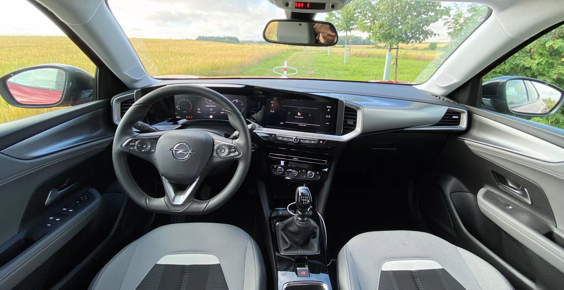 Pohľad na interiér zezadu sedadla automobilu Opel Mokka 2021 1.2 DI Turbo. Obsahuje trojramenný multifunkčný volant, digitálny rýchlomer a otáčkomer za volantom, infotainmentový dotykový displej na výšku, prepínače klimatizácie, manuálnu prevodovku s držiakmi nápojov, dvojfarebné športové predné sedadlá, zladené dverné panely s podrúčkou, viditeľné spätné zrkadlo, čiastočne otvorené okná a ďalšie ovládacie páčky pre stierače a ukazovatele smeru.
