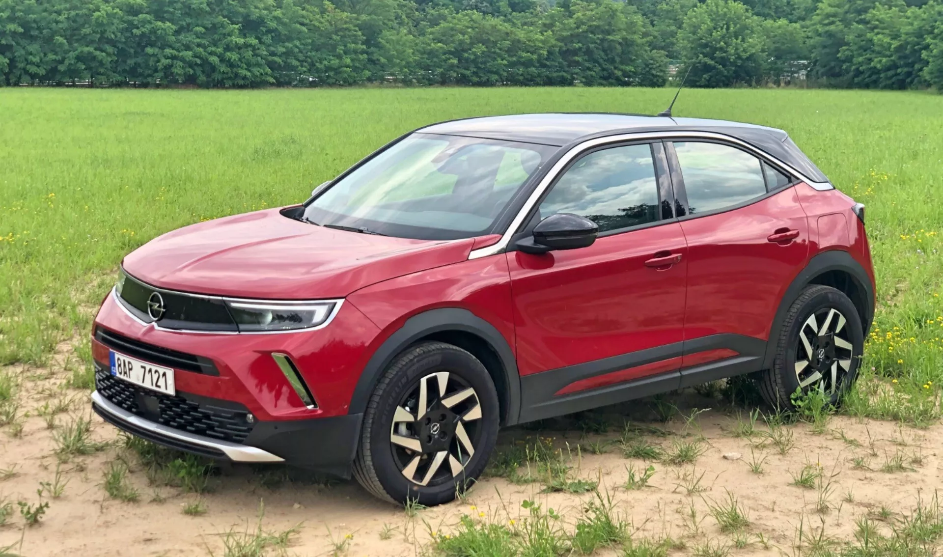 Obraz červeného SUV Opel Mokka 2021 zaparkovaného na trávnatej ploche s poľom v pozadí. Tento trojštvrťový predný pohľad zvýrazňuje mriežku, potenciálne naznačujúcu pokročilú technológiu pohonu, elegantné LED denné bežné svetlá, hmlové svetlá a jedinečné dvojfarebné kolesá z ľahkých zliatin. Viditeľné sú aj bočné zrkadlá a kľučky dverí v farbe karosérie, dynamická línia ramena, ochranná čierna spodná lišta a chrómové rámy okien, ktoré ukazujú moderné a luxusné dizajnové prvky vozidla.