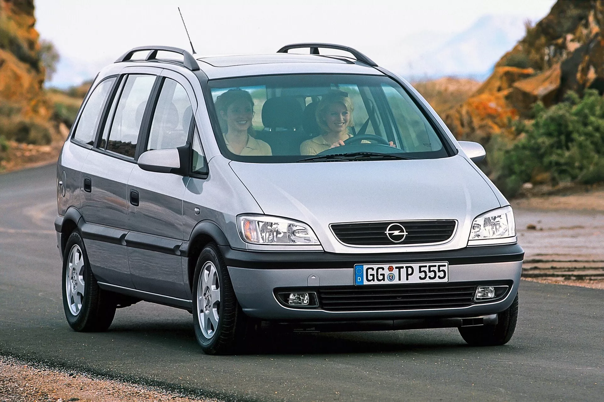 Fotografia kompaktného MPV Opel Zafira z roku 1999 z nízkej prednej trojštvrtinovej perspektívy. Obrázok zvýrazňuje mriežku chladiča so stredovým odznakom, veľké svetlomety pravdepodobne využívajúce halogénové svetelné zdroje, hladkú kapotu, predný nárazník s hmlovkami, hliníkové kolesá a pravú stranu vozidla s anténou na zadnej časti strechy. Vozidlo je na asfaltovanom povrchu s prírodnou krajinou v pozadí. Cez predné čelné sklo sú viditeľní dvaja cestujúci, čo odráža dizajn a priestrannosť interiéru vozidla.