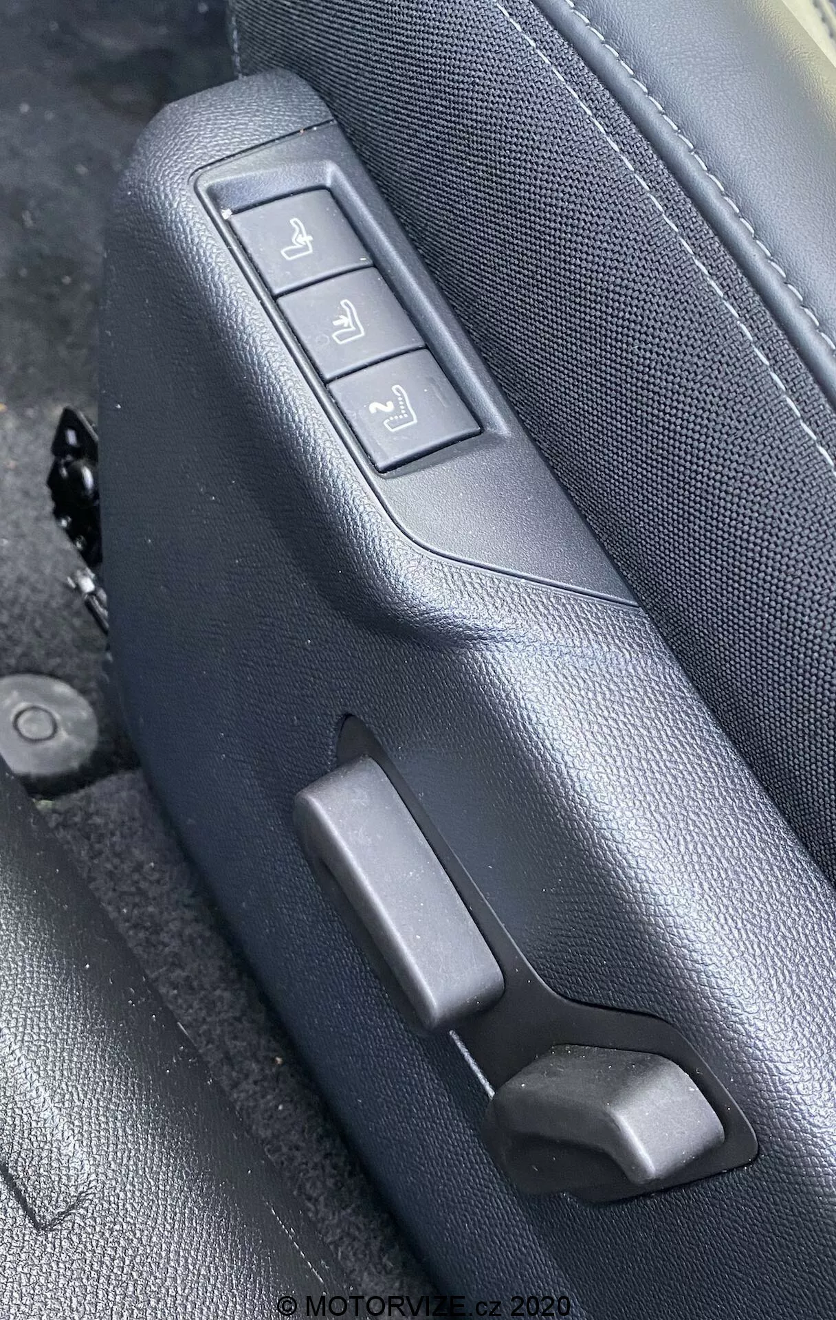 Detailný záber interiéru vozidla Peugeot 2008 1.2 PureTech 100 Active (2020) so zameraním na bočnú stranu čierneho alebo tmavosivého koženého sedadla s viditeľným prešívaním, z mierne nadhľadového uhla. Sedadlo je vybavené tromi elektronickými nastaviteľnými tlačidlami a ručnou pákou. Tlačidlá ovládajú pohyb dopredu/dozadu, náklon operadla a celkovú výšku sedadla, všetko na zodpovedajúcom textúrovanom paneli. Na obrázku je vodoznak s nápisom 'MOTORVIZE.cz 2020'.