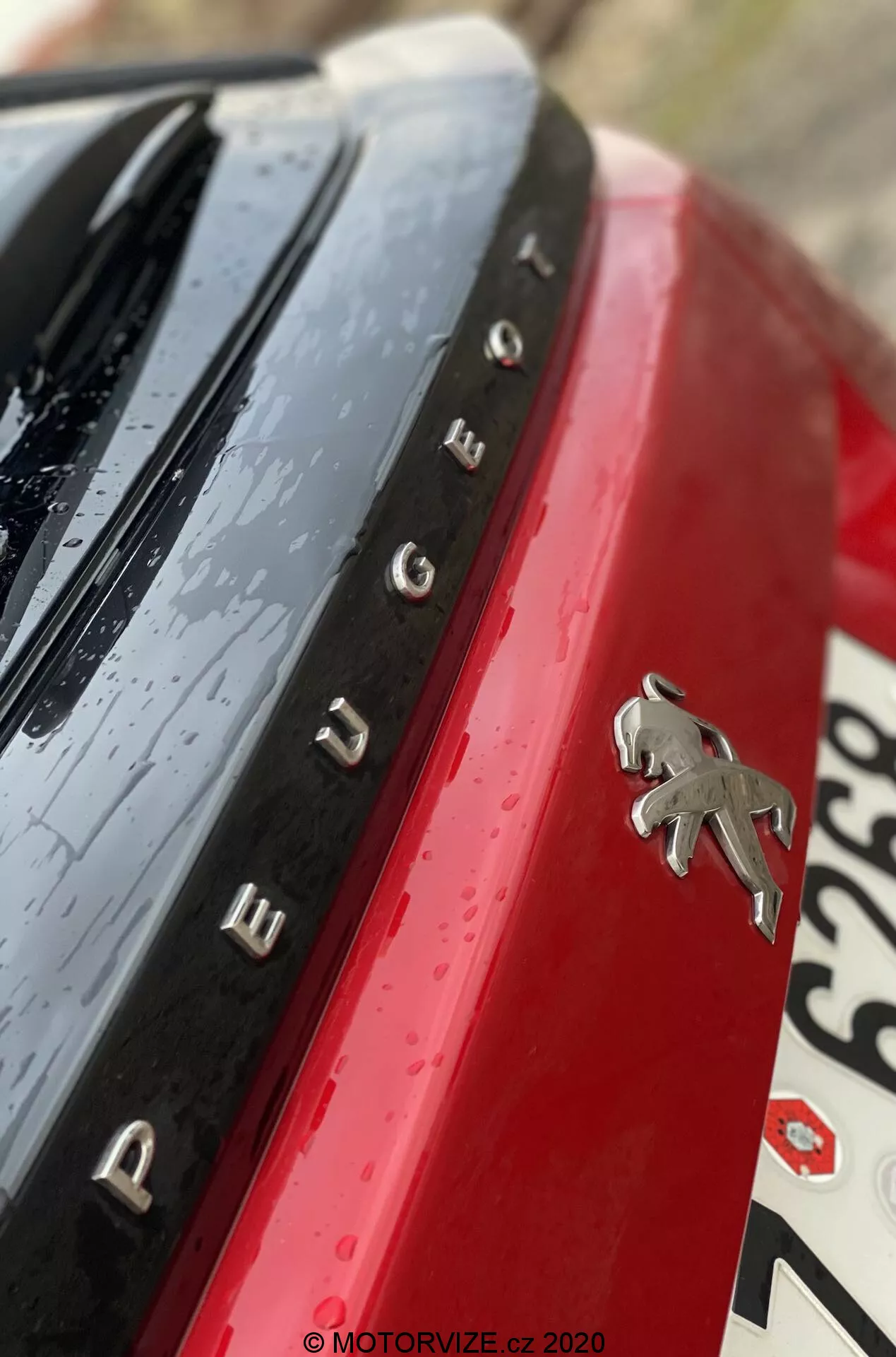 Detailný záber zadného emblému a označenia modelu Peugeot 2008 2020 (1.2 PureTech 100 Active). Snímka je urobená z neobvyklého diagonálneho uhla, ktorý zachytáva chrómové logo Peugeot na živom červenom paneli karosérie. Chrómové písmená vľavo od loga tvoria názov 'PEUGEOT'. Nad názvom je lesklý čierny povrch s kvapkami vody, čo naznačuje nedávny dážď alebo umývanie auta. Rozmazaná časť značky je viditeľná dole a vpravo. Plytká hĺbka ostrosti zaostruje emblém a názov, ostatné prvky sú rozmazané pre umelecký nádych.