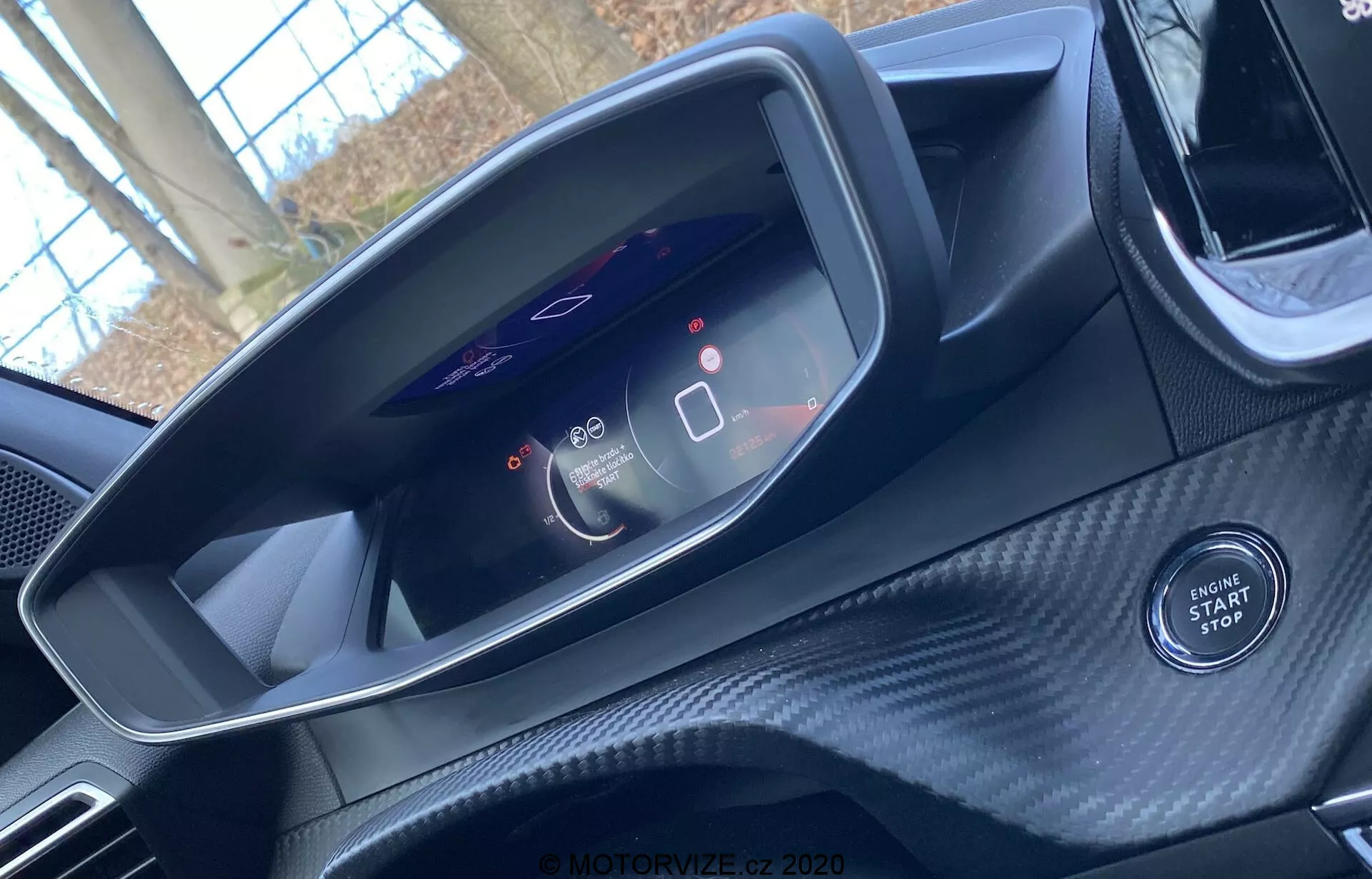 Pohľad na interiér Peugeot 2008 2020 z pohľadu vodiča. Obrázok sa zameriava na moderný digitálny prístrojový panel zobrazujúci tachometer na 0 a merač spotreby energie. Zobrazuje tiež ikony pokročilých asistenčných systémov vodiča. Tlačidlo štartu/stopu motora je viditeľné pod prístrojovým panelom, čo naznačuje bezkľúčový zapaľovací systém auta. Oblasť okolia má textúru podobnú uhlíkovým vláknam, čo naznačuje športový alebo luxusný interiér. Horná časť volantu je len ťažko viditeľná.