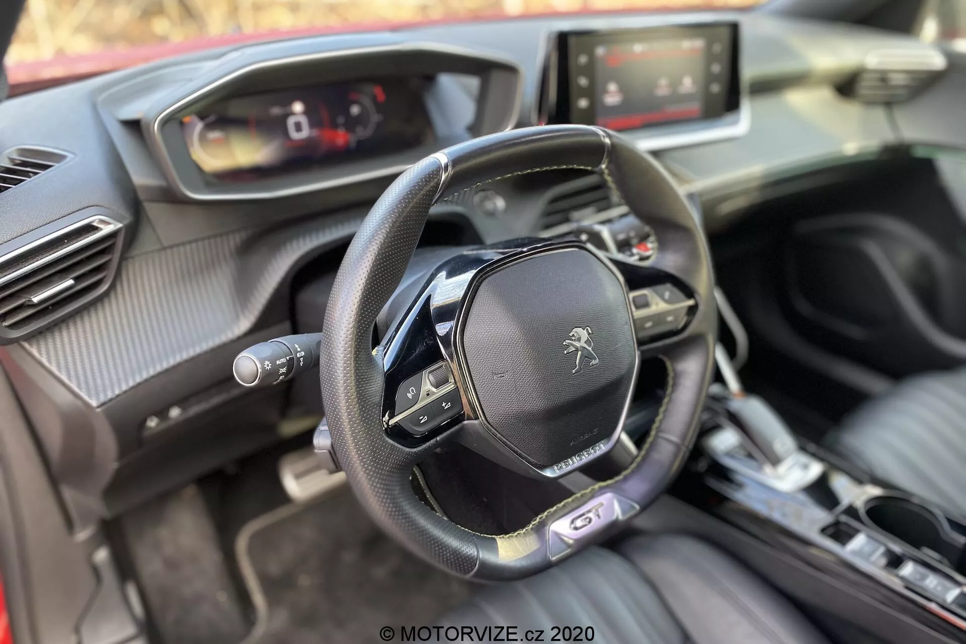 Interiér vozidla Peugeot 2008 1.2 PureTech 100 Active z roku 2020 z pohľadu predného spolujazdca zameraného na kokpit vodiča. Obrázok ukazuje rovné spodné volantu s viacerými ovládacími tlačidlami a logom leva Peugeot, digitálny prístrojový panel, dotykový infotainment systém, centrálne ovládanie automatickej prevodovky, tlačidlo štart/stop zapaľovania, vzduchové prieduchy s chrómovanými akcentmi a zmes mäkkých materiálov vrátane kože a textúr podobných uhlíkovým vláknam.