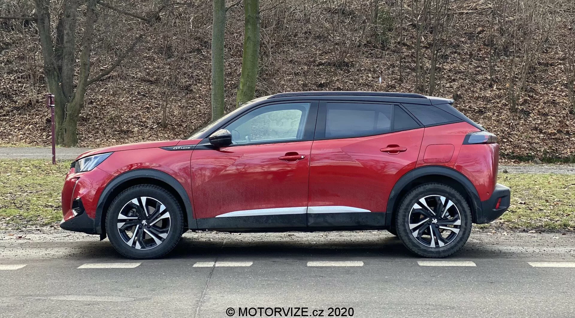 Červené športové úžitkové vozidlo (SUV) Peugeot 2008 2020 1.2 PureTech 100 Active s moderným a elegantným dizajnom, so zvažujúcou sa líniou strechy, vysokým pásom so zužujúcimi sa oknami, aerodynamickými kľučkami dverí, zrkadlá s integrovanými smerovkami, veľké viacramenné zliatinové kolesá, nízkoprofilové pneumatiky, čierne plastové lemovanie pre robustný vzhľad, lesklá červená farba a tónované zadné okná, zachytené v profilovom pohľade zaparkované v vonkajšom prostredí s bezlistými stromami a trávnatým pozadím.