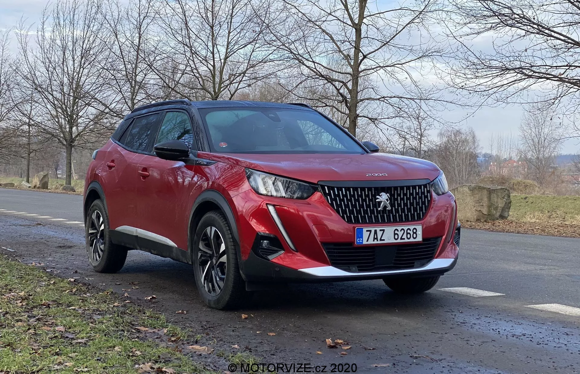 Červené SUV Peugeot 2008 2020 (Peugeot-2008-1-2-Puretech-100-Active) z predného trojštvrťového pohľadu, zvýrazňujúci prednú mriežku s výrazným emblémom značky, elegantnými LED svetlami, prednými parkovacími senzormi a hmlovými svetlami. Pohľad zboku odhaľuje viacpaprskové hliníkové kolesá, vyššiu svetlú výšku, dynamické línie karosérie, integrované smerovky na bočných zrkadlách a čierne plastové obloženie. Položené pod ľahkým uhlom na strane cesty s prírodnou, vidieckou krajinou v pozadí, čo naznačuje neskorú jeseň alebo skorú jar. Nízky uhol kamery pre silný a impozantný pohľad.