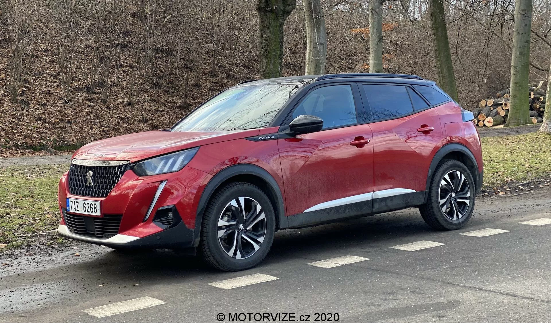 Bočný a predný pohľad na moderné crossover SUV Peugeot 2008 1.2 PureTech 100 Active z roku 2020, zdôrazňujúci jeho výraznú masku chladiča, elegantné svetlomety, hliníkové kolesá a dynamické línie karosérie. Vozidlo, ozdobené červeným metalickým lakom s tónovanými zadnými oknami, je zobrazené zaparkované na bočnej ceste s viditeľnými strešnými lištami, čo naznačuje mestskú a ľahkú terénnu schopnosť. Obrázok pochádza z MOTORVIZE.cz 2020.