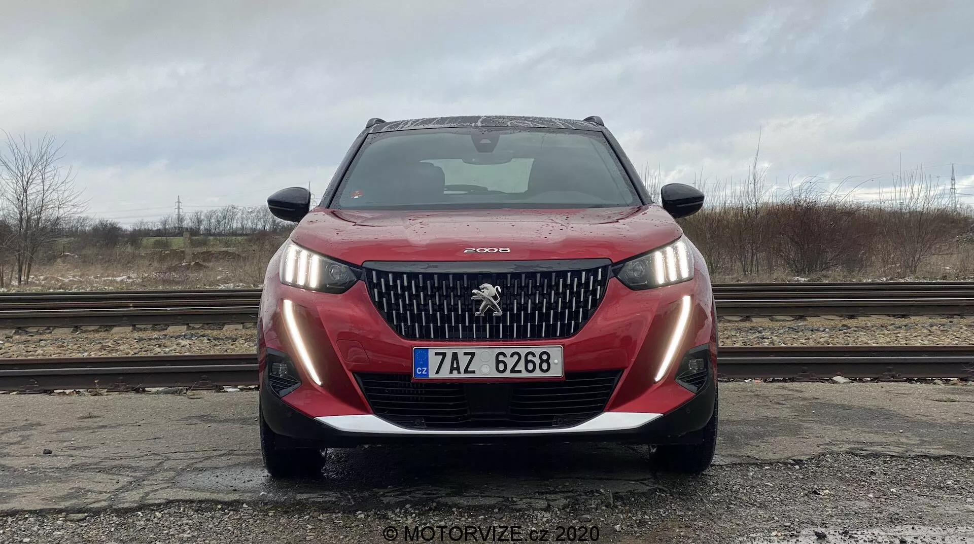 Pohľad spredu na moderný model Peugeot 2008 2020, zachytený z nízkeho uhla. Obrázok zobrazuje prednú masku vozidla, charakteristické logo leva, LED svetlomety a nárazník. Maska má vertikálne lamely typické pre dizajnový jazyk Peugeotu. Uhol zvýrazňuje šírku a prítomnosť vozidla na ceste. Nárazník obsahuje lakované a čierne prvky, čo naznačuje športový dizajn, s viditeľnými parkovacími senzormi a registračnou značkou v strede. Scéna je nasnímaná za zamračeného dňa s rozmazaným pozadím a holými stromami v diaľke.