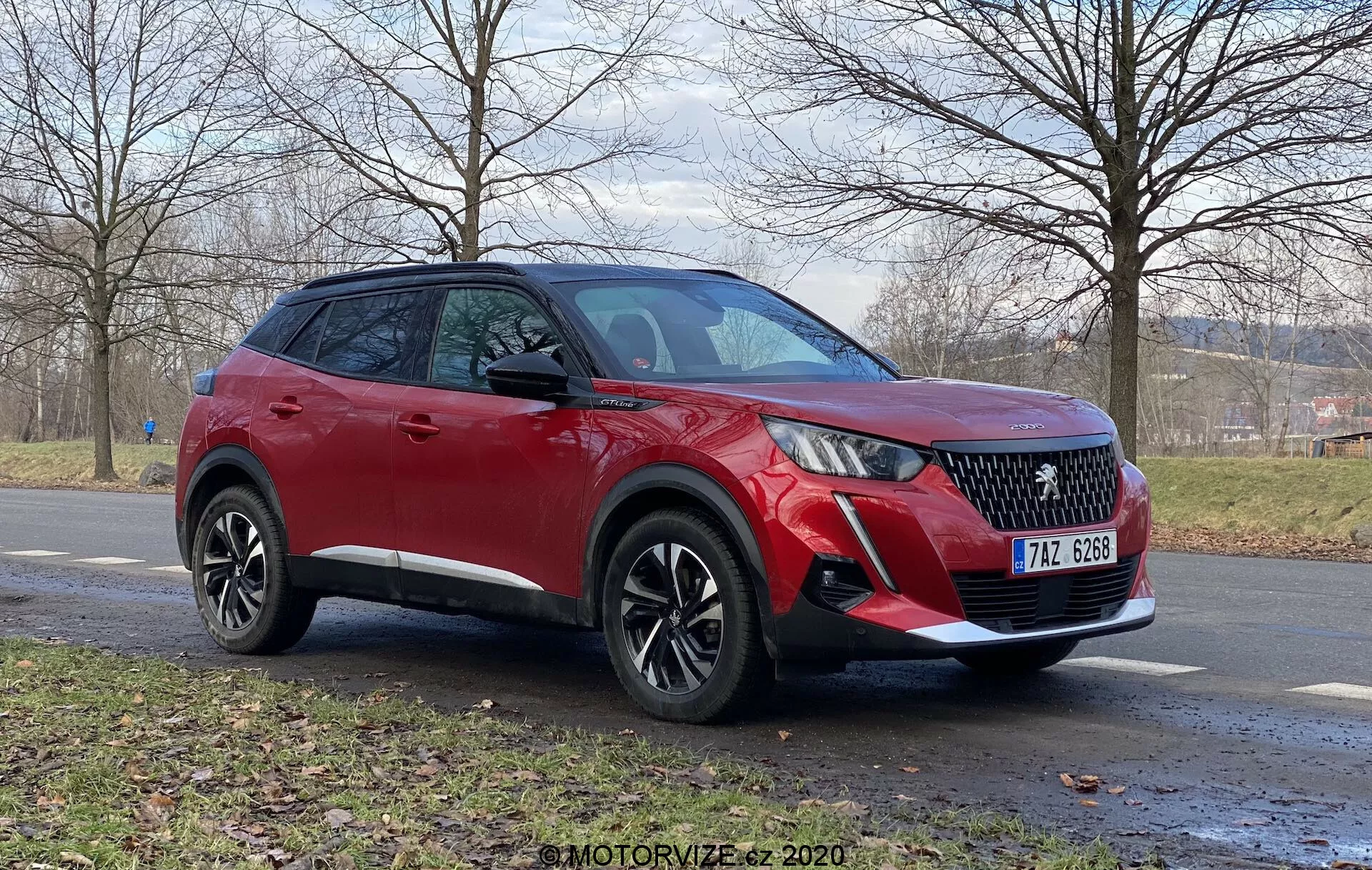 Trojštvrťový predný pohľad na červený Peugeot 2008 2020 (1.2 PureTech 100 Active) crossover SUV na asfaltovej ceste. Viditeľné vlastnosti zahŕňajú LED denné svetlá, mriežku chladiča s diamantovým vzorom, predný nárazník s hmlovými svetlami a možné parkovacie senzory, liatinové kolesá s viacerými lúčmi a kameru pod bočným zrkadlom vo farbe karosérie, všetko pod zatiahnutou oblohou. V pozadí je trávnatá plocha a strommy bez lístia, čo naznačuje neskorú jeseň alebo zimu.