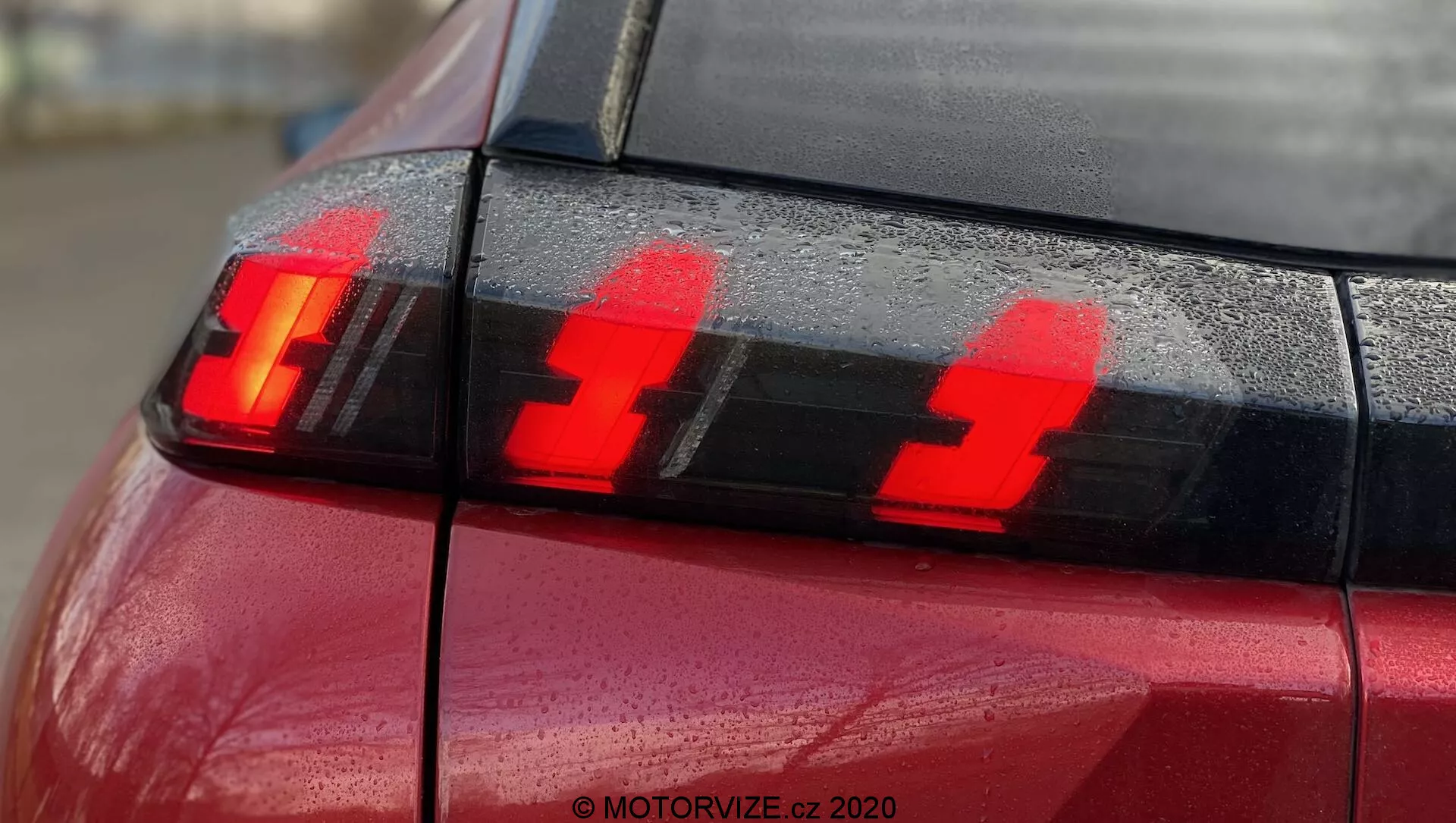 Záber na zadnú časť červeného Peugeota 2008 2020 (PureTech 100 Active) so zameraním na zostavu zadných svetiel so svietiacimi brzdovými svetlami. Fotografia, urobená zo zadného rohu mierne nabok, zvýrazňuje tri zvislé LED svetelné prvky a kvapky vody na povrchu, zdôrazňujúce moderný dizajn osvetlenia vozidiel.