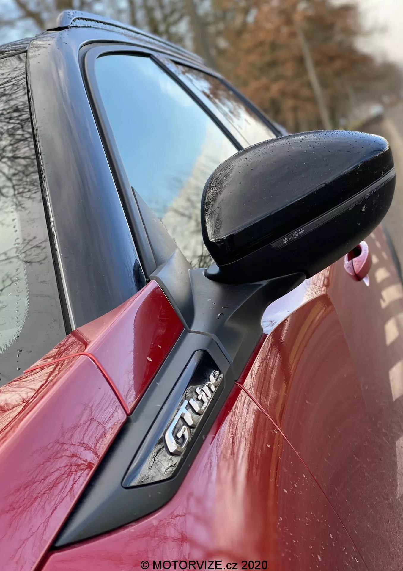 Detailný záber na bok červeného vozu Peugeot 2008 2020, zameraný na čierne spätné zrkadlo na strane vodiča, upevnené na dverách čiernou podporou. Čiastočne viditeľný stĺpik A s emblémom 'GT Line', ktorý zvýrazňuje aerodynamické línie vozidla a športové vybavenie. Vodotlač od MOTORVIZE.cz 2020.
