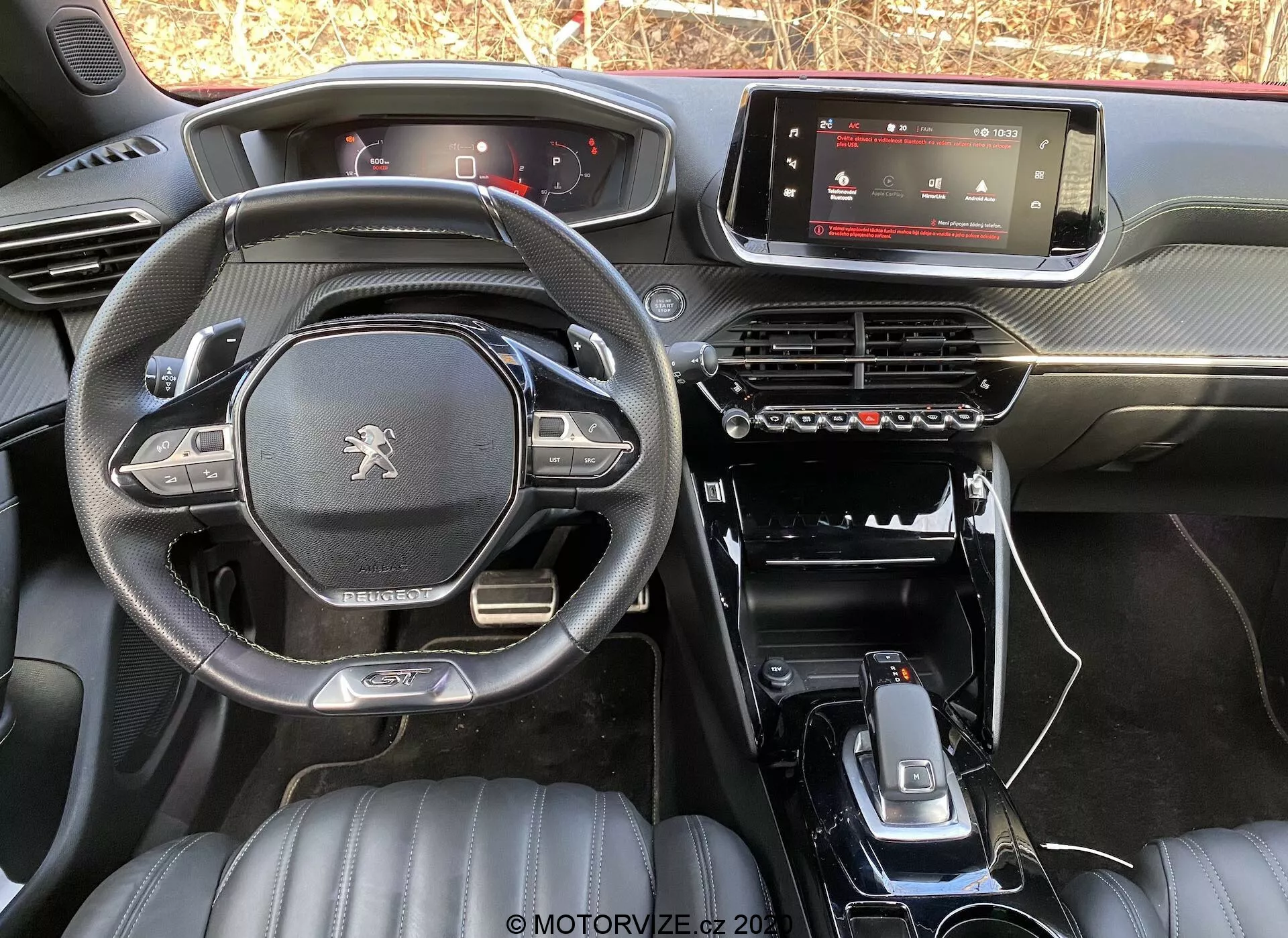 Pohľad na interiér vozidla Peugeot 2008 1.2 PureTech 100 Active 2020 zo zadného sedadla, ktorý ukazuje palubnú dosku, digitálny infotainment displej, panel klimatizácie, minimalistickú stredovú konzolu s radiacou pákou, držiakmi na nápoje a ďalšími ovládacími tlačidlami. Volant s plochým dnom má logo Peugeot a multifunkčné ovládacie prvky. Predné kožené sedadlá s prešívaným vzorom naznačujú vyššiu úroveň výbavy. Prirodzené svetlo zvýrazňuje dizajn a materiály továrenského modelu.