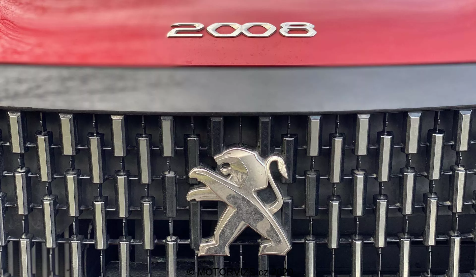 Zblízka pohľad na prednú mriežku automobilu Peugeot 2008 1.2 PureTech 100 Active 2020, ktorý zobrazuje charakteristický dizajn s výraznou červenou kapotou, zložité usporiadanie čiernych vertikálnych a horizontálnych lamiel a lesklý strieborný emblém leva v strede. Fotografia sa zameriava na estetické a brandingové prvky a poskytuje jasnú vizualizáciu detailov mriežky.