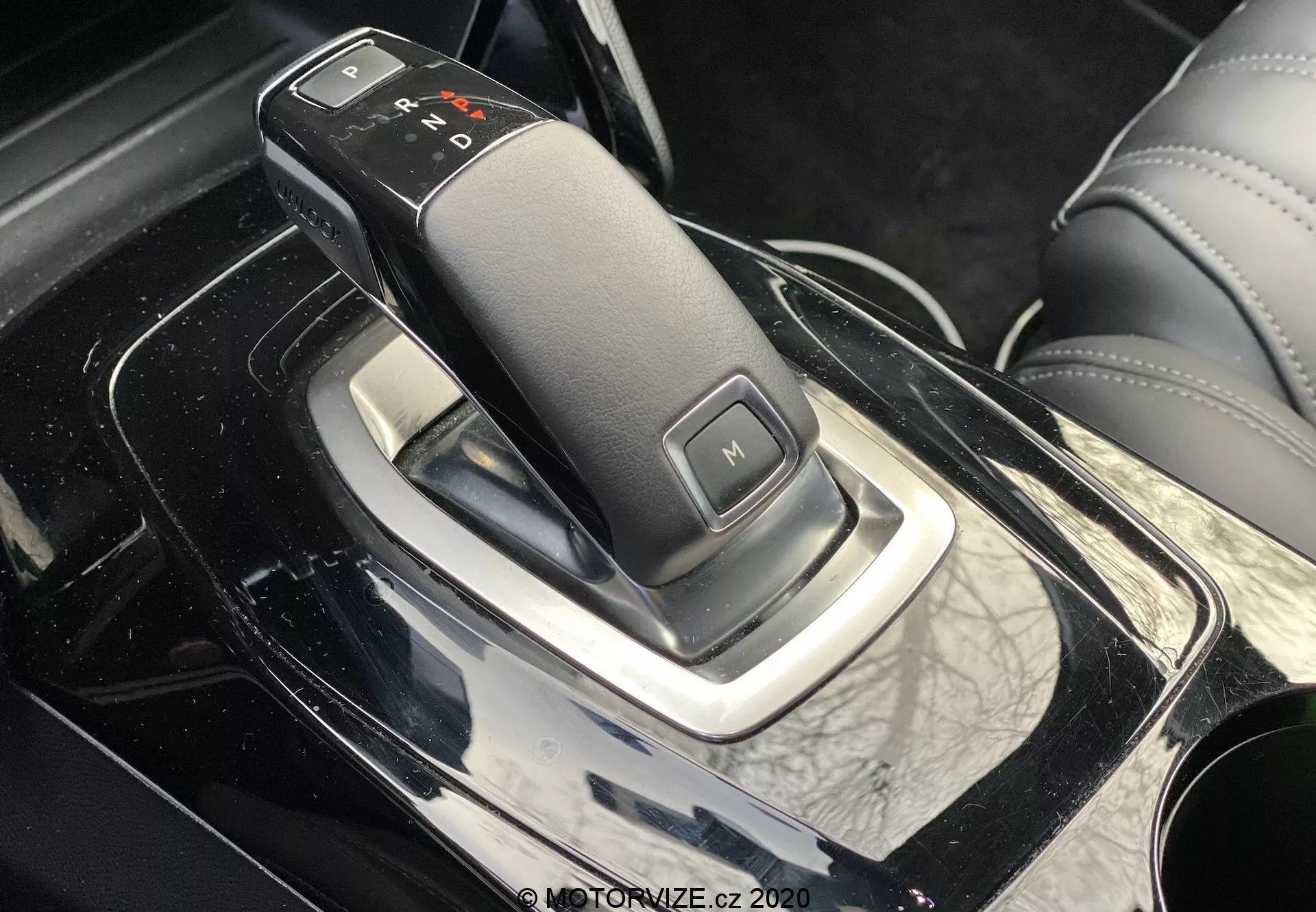 Blízky, bočný pohľad na interiér Peugeot 2008 2020 s elegantnou pákou prevodovky, s automatickou prevodovkou označenou 'P', 'R', 'N', 'D' a 'M' pre manuálny režim, na lesklej, čiernej stredovej konzole s chrómovými akcentmi a koženým okolím, čo naznačuje luxusný interiér auta.