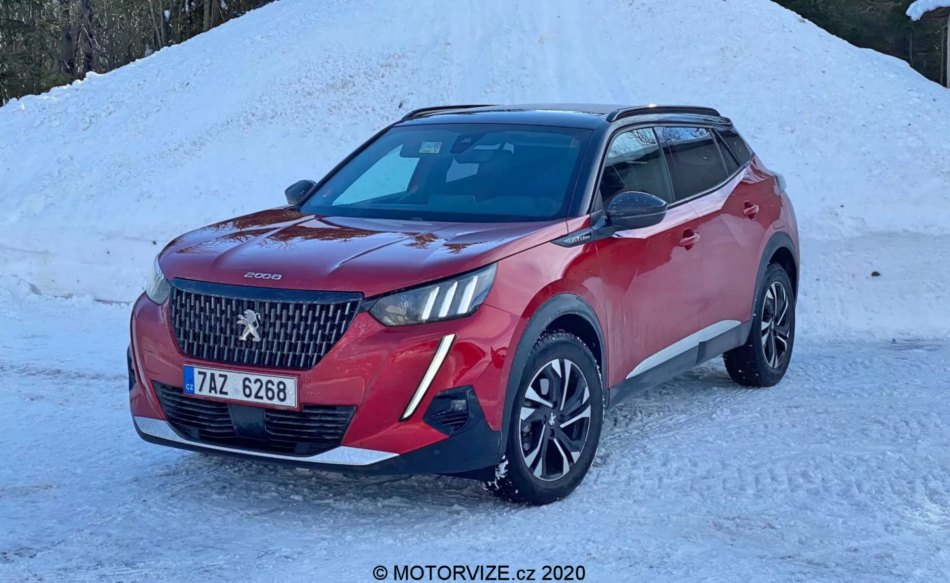 Obrázok červeného SUV Peugeot 2008 2020 (peugeot-2008-1-2-puretech-100-active) zaparkovaného na zasneženej zemi. Moderné vozidlo je zobrazené pod uhlom troch štvrtín zepredu, zvýrazňujúcou jeho výraznú mriežku s veľkým logo v strede a diamantovým vzorom. Viditeľné sú štýlové LED svetlomety, dvojfarebná karoséria s čiernymi krytmi zrkadiel a strechou, viaclúčové zliatinové kolesá a hmlové svetlá alebo doplnkové osvetlenie na prednom nárazníku. Auto má predné parkovacie senzory, kamerový alebo senzorový modul na čelnom skle pre asistenčnú technológiu pre vodiča a strešnú anténu v tvare žraločej plutvy naznačujúcu funkciu GPS alebo satelitného rádia. Scéna naznačuje zimné prostredie s jasným prirodzeným svetlom zvýrazňujúcim detaily auta.
