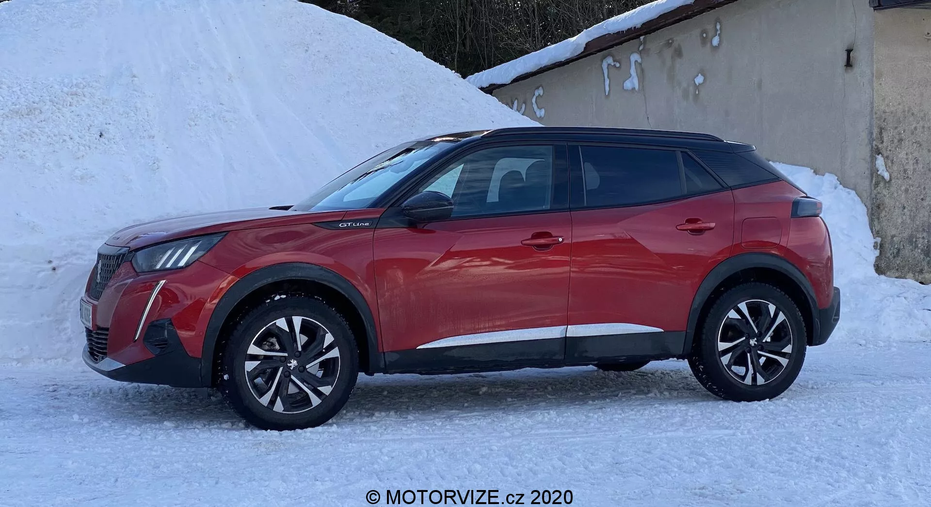 Bočný profil moderného SUV Peugeot 2008 z roku 2020 v živej a lesklej červenej farbe. Vozidlo je umiestnené na zasneženom povrchu, čo naznačuje vhodnosť pre zimné podmienky. Dynamický a uhlový dizajn zahŕňa prominentné blatníky, ostré línie karosérie, sklonenú líniu strechy, hliníkové kolesá a bočné zrkadlá v farbe karosérie s pokročilými funkciami ako elektrické nastavenie alebo sledovanie mŕtveho uhla. Štvordverové usporiadanie s tmavými oknami, čiernym dolným lemovaním a zložitými diskami zvyšuje športovú aj robustnú estetiku.