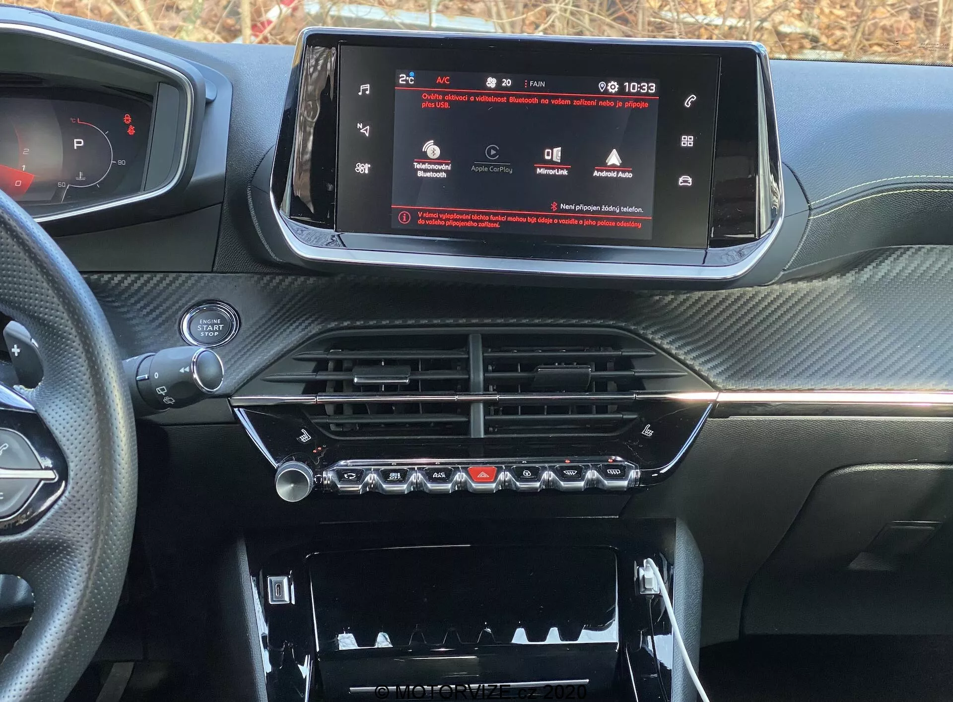 Detailný pohľad na interiérovú palubnú dosku moderného auta, konkrétne na model Peugeot 2008 2020 (peugeot-2008-1-2-puretech-100-active). Obraz je urobený z miesta vodiča alebo predného spolujazdca a sústreďuje sa na centrálnu konzolu a infotainment systém. Širokouhlý dotykový displej zobrazuje menu nastavenia vozidla, čo naznačuje pokročilú technológiu. Nižšie sú chrómované vetracie otvory a ovládanie klímy s fyzickými tlačidlami a otočnými ovládačmi. Nižšie je viditeľný priestor s krytom. Palubná doska má textúrovaný, karbonový povrch, čo dodáva športový a luxusný vzhľad.
