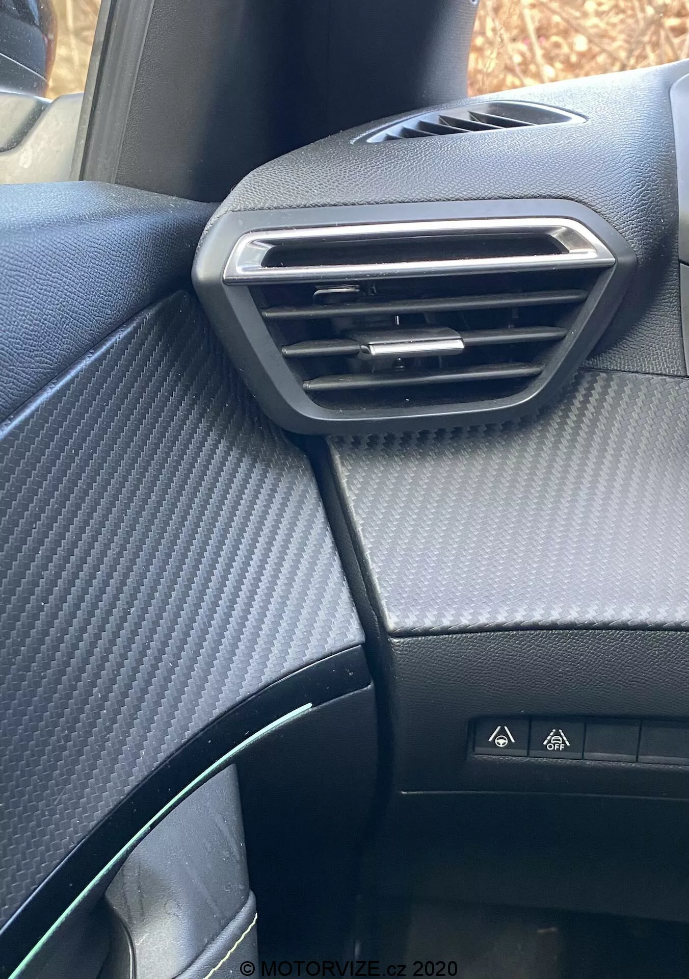 Detailný pohľad na palubnú dosku na strane spolujazdca v interiéri Peugeot 2008 2020, zvýrazňujúci strieborný nastaviteľný vetrací otvor, hornú palubnú dosku s textúrou karbónových vlákien, tlačidlo výstražného svetla, čiastočný volant a čierne sedadlo s bodkovaným vzorom.