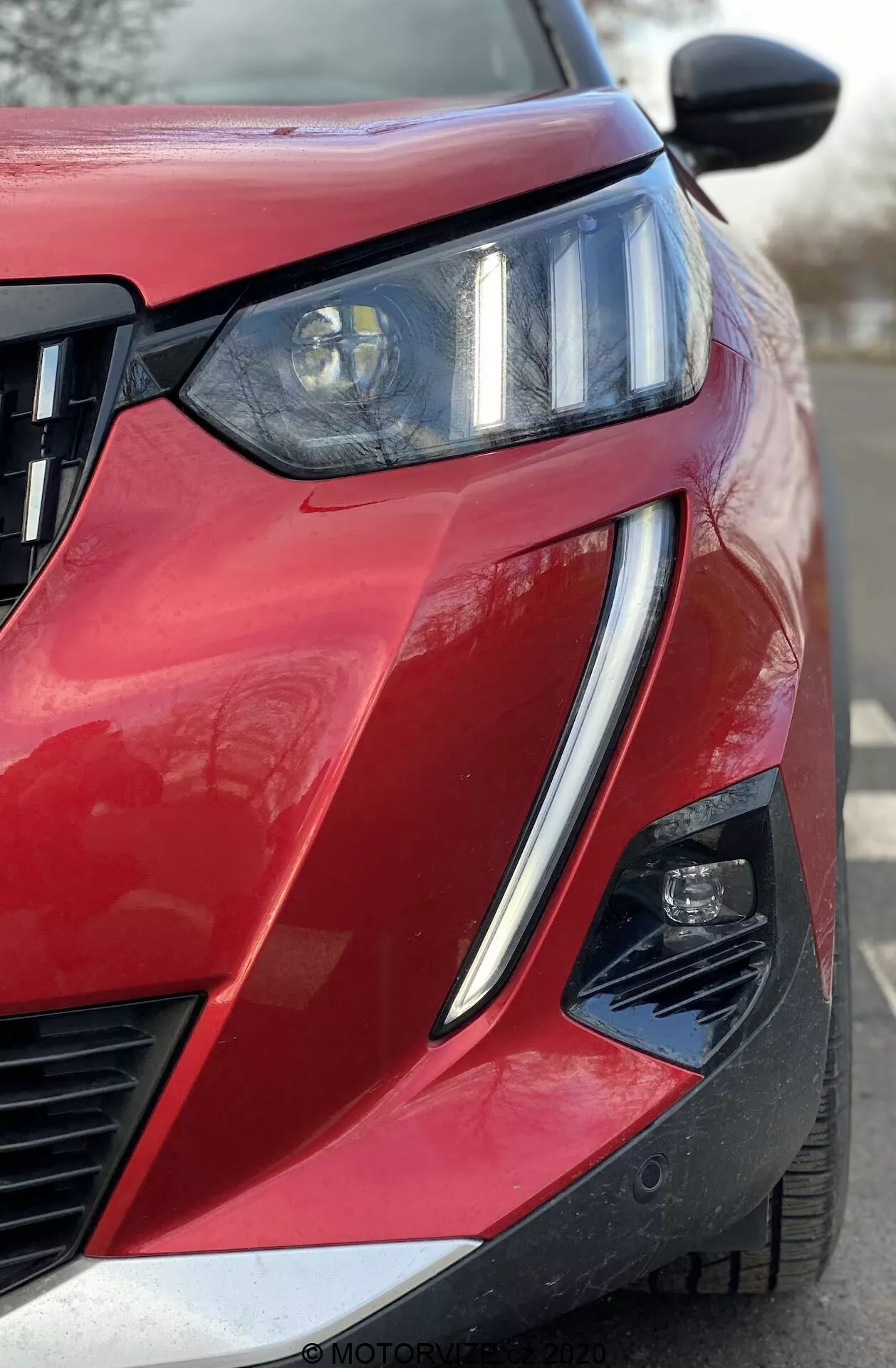 Detailný uhlový predný pohľad na červený Peugeot 2008 1.2 PureTech 100 Active z roku 2020, zameraný na predný ľavý kvadrant. Výrazný reflektor s projektorom a integrovanými LED DRL svetlami, horizontálna mriežka chladiča a čiastočne viditeľné sekundárne osvetľovacie teleso v uhlovej výreze nárazníka. Lesklý lak s metalickým povrchom, ostrá línia karosérie, zvýšený obrys kapoty, čierne spätné zrkadlo s pravdepodobne integrovanými smerovkami alebo kamerou pre monitorovanie slepého uhlu a výrez v dolnom rohu nárazníka naznačujúci pokročilé asistenčné systémy pre vodiča.