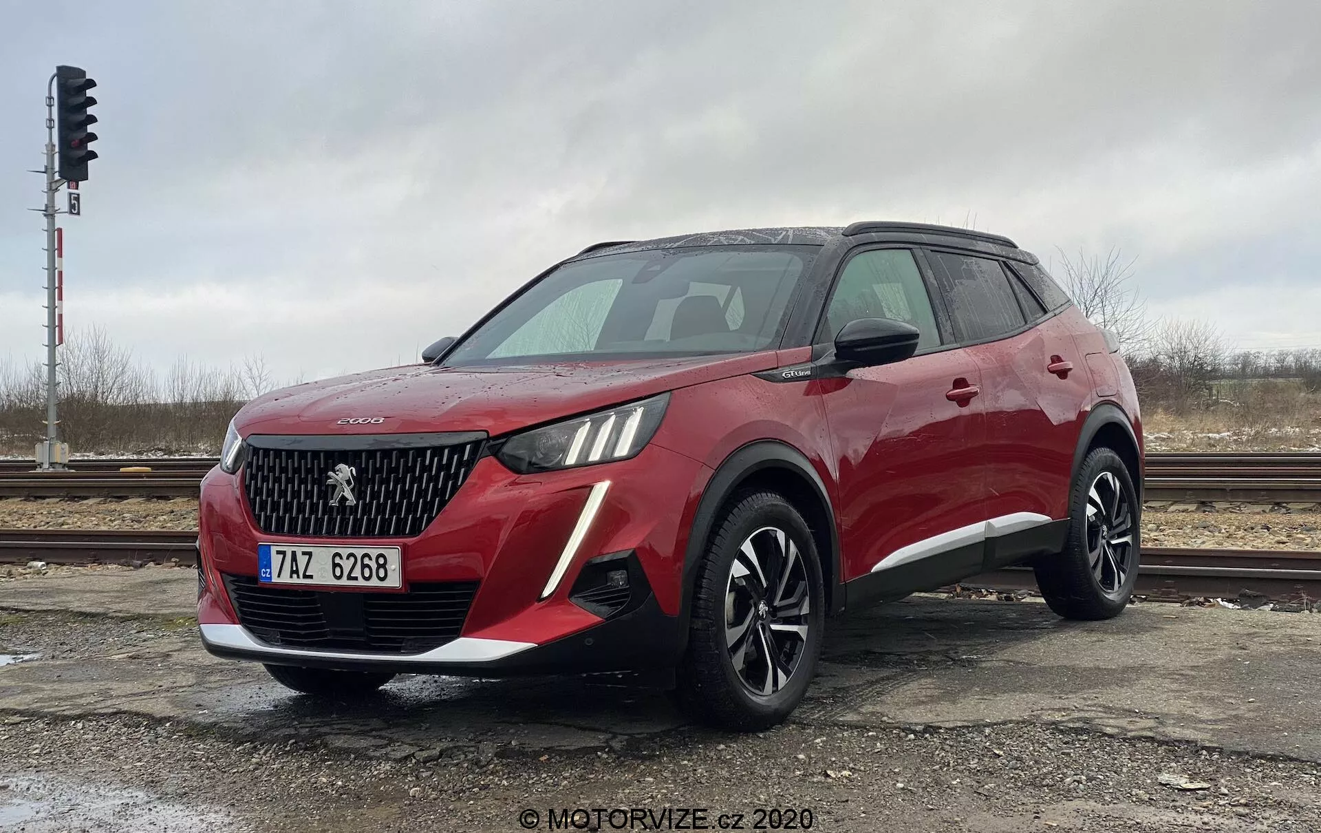 Trojštvťový predný pohľad na moderné, červené crossover SUV Peugeot 2008 1.2 PureTech 100 Active. Nízky uhol záberu zdôrazňuje výšku a postoj vozidla. Predná časť ukazuje výraznú mriežku s diamantovým vzorom, emblém pravdepodobne reprezentujúci výrobný znak a charakteristické svetlomety s LED denným osvetlením. Predný nárazník obsahuje hmlovky alebo prívody vzduchu. Bočný profil odhaľuje aerodynamické línie, veľké viacramenné liate kolesá, strešné nosiče a bočné zrkadlá s integrovanými smerovkami. Obrázok zachytáva dizajn a technologické prvky SUV za oblačného dňa, zaparkované pri železničných tratiach.