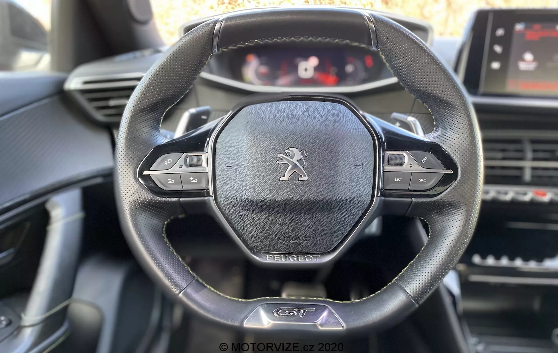Interiér vozidla Peugeot 2008 2020 zameraný na volant a palubnú dosku z pohľadu vodiča. Hlavné prvky zahŕňajú kožený trojramenný volant s multifunkčnými tlačidlami, emblém leva značky Peugeot a konfigurovateľný digitálny prístrojový panel. Viditeľné prvky zahŕňajú dotykovú obrazovku infotainmentu, vetracie otvory a ovládacie prvky na stredovom paneli s jemným denným osvetlením zdôrazňujúcim detaily volantu.