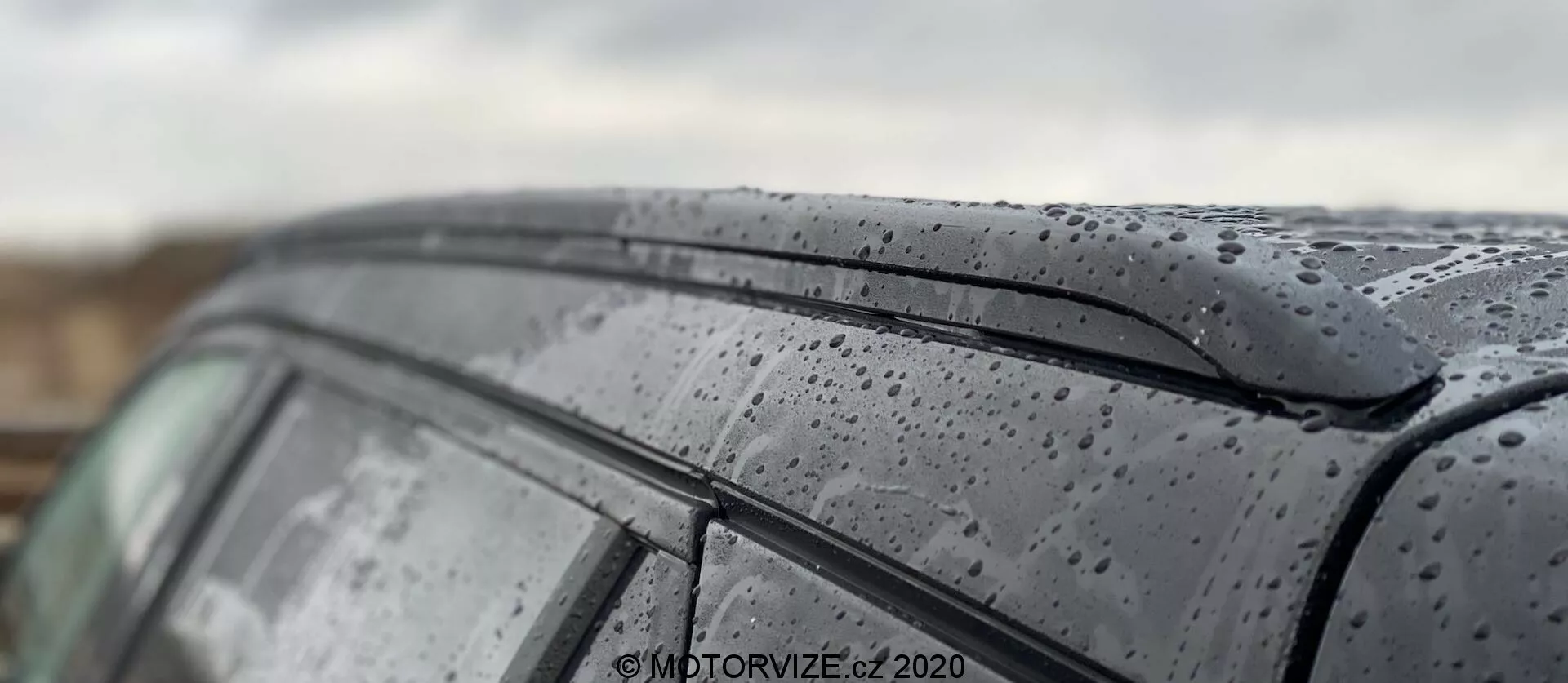 Detailný záber na strechu a bočné okno modelu Peugeot 2008 1.2 PureTech 100 Active 2020, zdôrazňujúci kvapky dažďa na elegantnom povrchu. Snímka zachytáva tmavý lesklý lak s kvapkami dažďa a minimalistický dizajn horného okraja bočného okna. Plytká hĺbka ostrosti zdôrazňuje textúru exteriéru vozidla, zatiaľ čo nejasne zobrazuje pozadie a časti strechy, čo predstavuje umelecké zobrazenie auta v mokrých podmienkach.