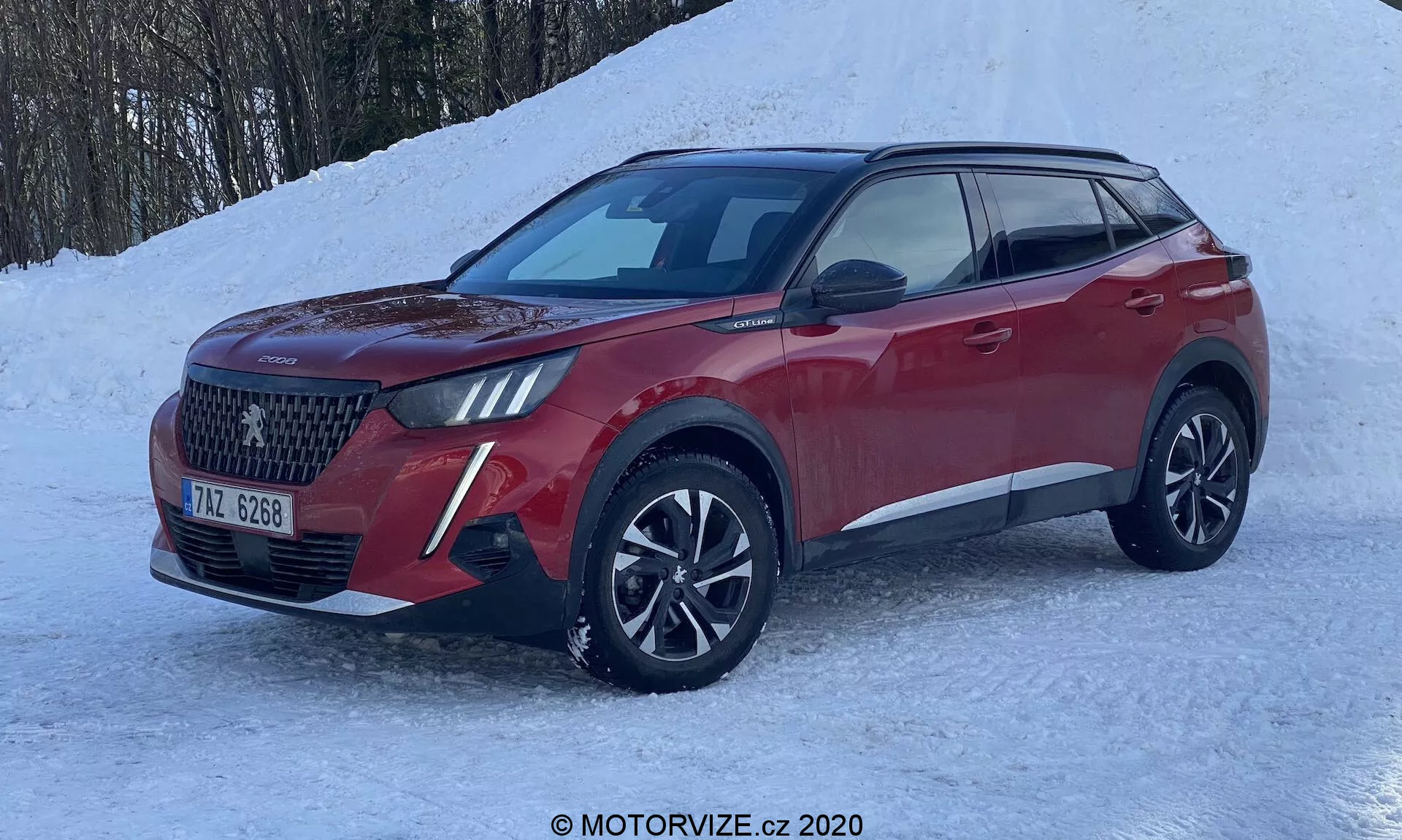 Moderné SUV Peugeot 2008 1.2 PureTech 100 Active 2020 v trojštvrťovom pohľade spredu, zvýrazňujúce veľkú mriežku s diamantovým vzorom, LED svetlomety s dennými svetlami, viacpaprskové zliatinové kolesá a sochársku kovovú červenú karosériu. Umienstnené na zasneženej zemi, naznačujúce zimné jazdné schopnosti ako pohon všetkých kolies.