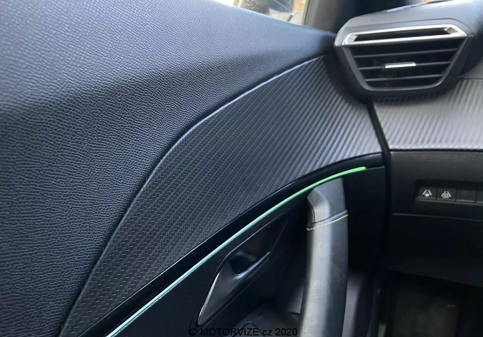 Detailný pohľad na interiérovú palubnú dosku a detaily na strane spolujazdca vozidla Peugeot 2008 2020, zameriavajúci sa na textúrovanú čiernu syntetiku pripomínajúcu karbónové vlákna, viditeľný nastaviteľný ventilátor, čiastočne otvorené dvere so zeleným odrazom a chrómovým akcentom, kľučky dverí, drôt s nabíjacím konektorom, kabínové svetlo a neidentifikované tlačidlo, zachytené s plytkou hĺbkou ostrosti a prirodzeným osvetlením.