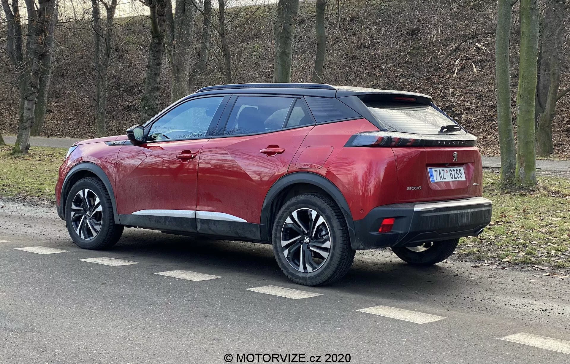 Červené SUV Peugeot 2008 1.2 PureTech 100 Active zachytené z trojštvrťového zadného uhla, zobrazujúce moderné obalové LED zadné svetlá, vysokú svetlú výšku, päťramenné hliníkové kolesá, klesajúcu líniu strechy, ostrú líniu karosérie, viditeľné kľučky dverí, automaticky skladacie bočné zrkadlá so zabudovanými smerovkami, chrómované lišty okien a antennu žraločia plutva. Vozidlo je zaparkované na asfaltovanom povrchu pri parku alebo lesnej oblasti, zvýraznené prirodzeným osvetlením.