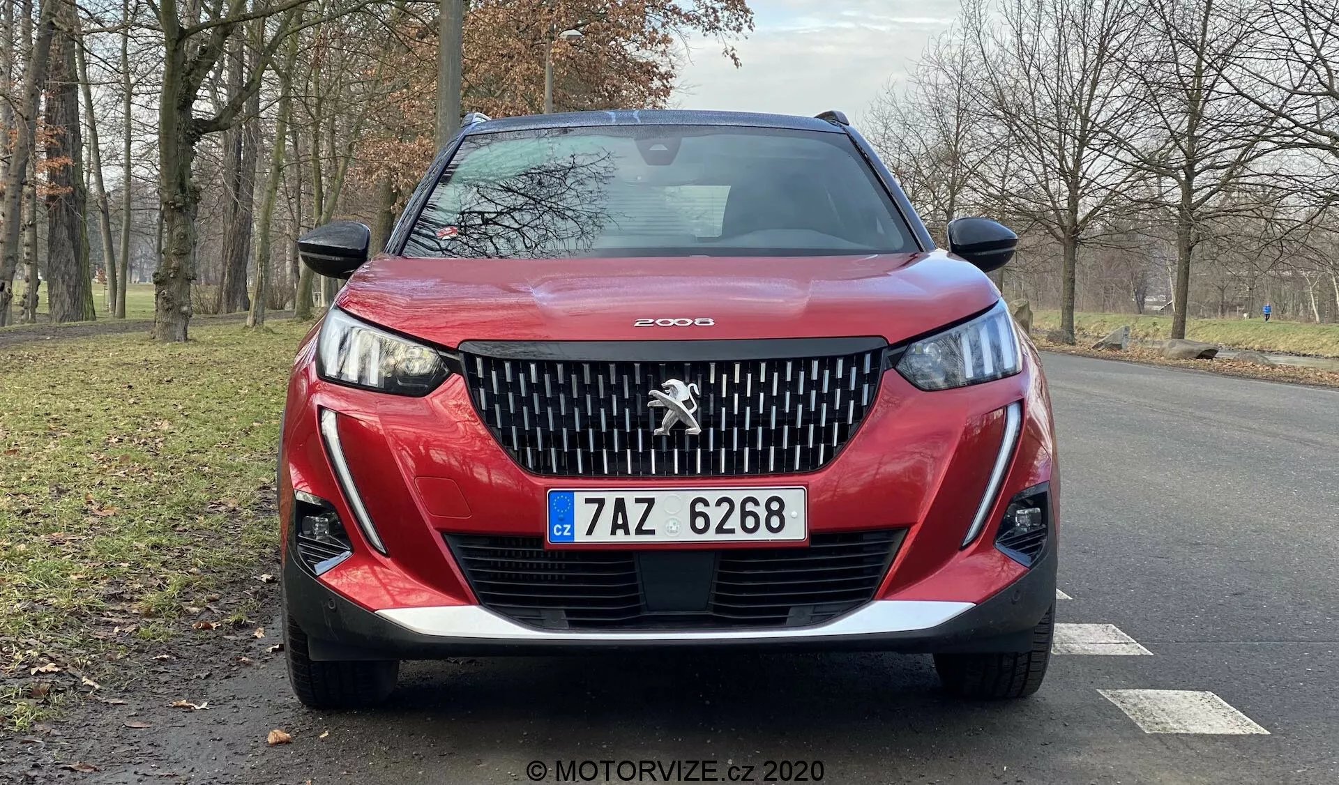 Blízky čelný pohľad na červený Peugeot 2008 2020 (Peugeot-2008-1-2-PureTech-100-Active) zobrazujúci detailnú prednú mriežku s výrazným logom leva Peugeot, agresívny dizajn svetlometov s dennými LED svetlami v tvare 'tesákov', tvarovaný dolný nárazník a českú poznávaciu značku. Obrys kapoty pridáva dynamiku a obrázok zachytáva čiastočný pohľad na zliatinové kolesá a spätné zrkadlá na suchej asfaltovej ceste s bezlistými stromami a trávnatou plochou v pozadí, čo naznačuje pokojné, prírodné prostredie, možno na jeseň alebo v zime.